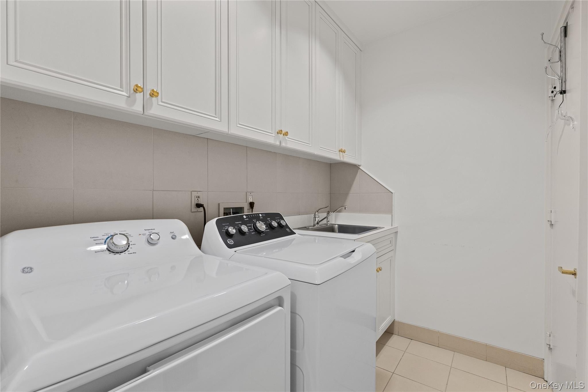 Laundry area featuring white shaker-style cabinetry, a stainless steel utility sink, tile backsplash, and light-toned tile flooring