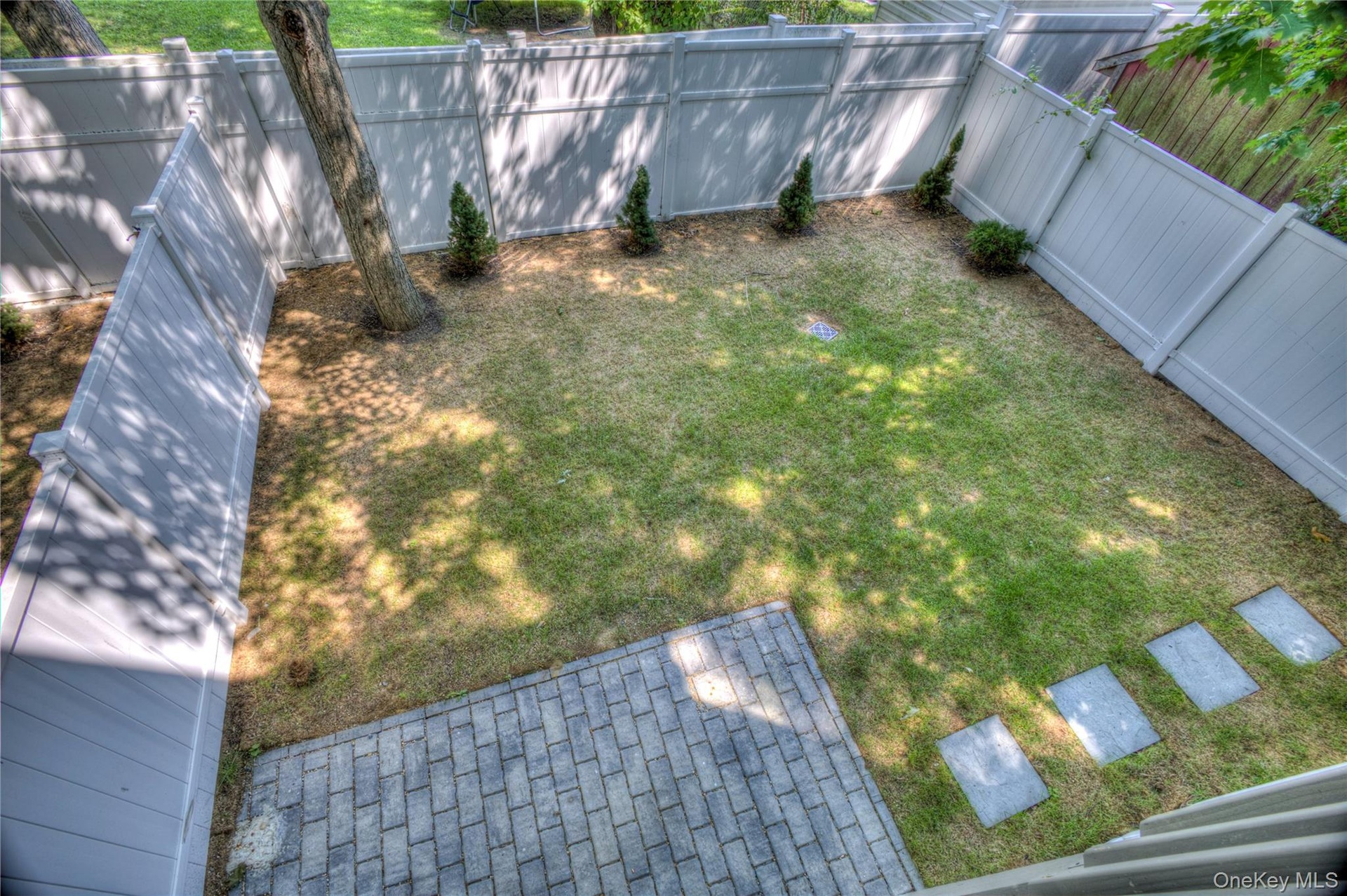 Rear yard featuring a full-height vinyl privacy fence, a grey stone paver patio, a manicured lawn, stone stepping stones, and mature trees