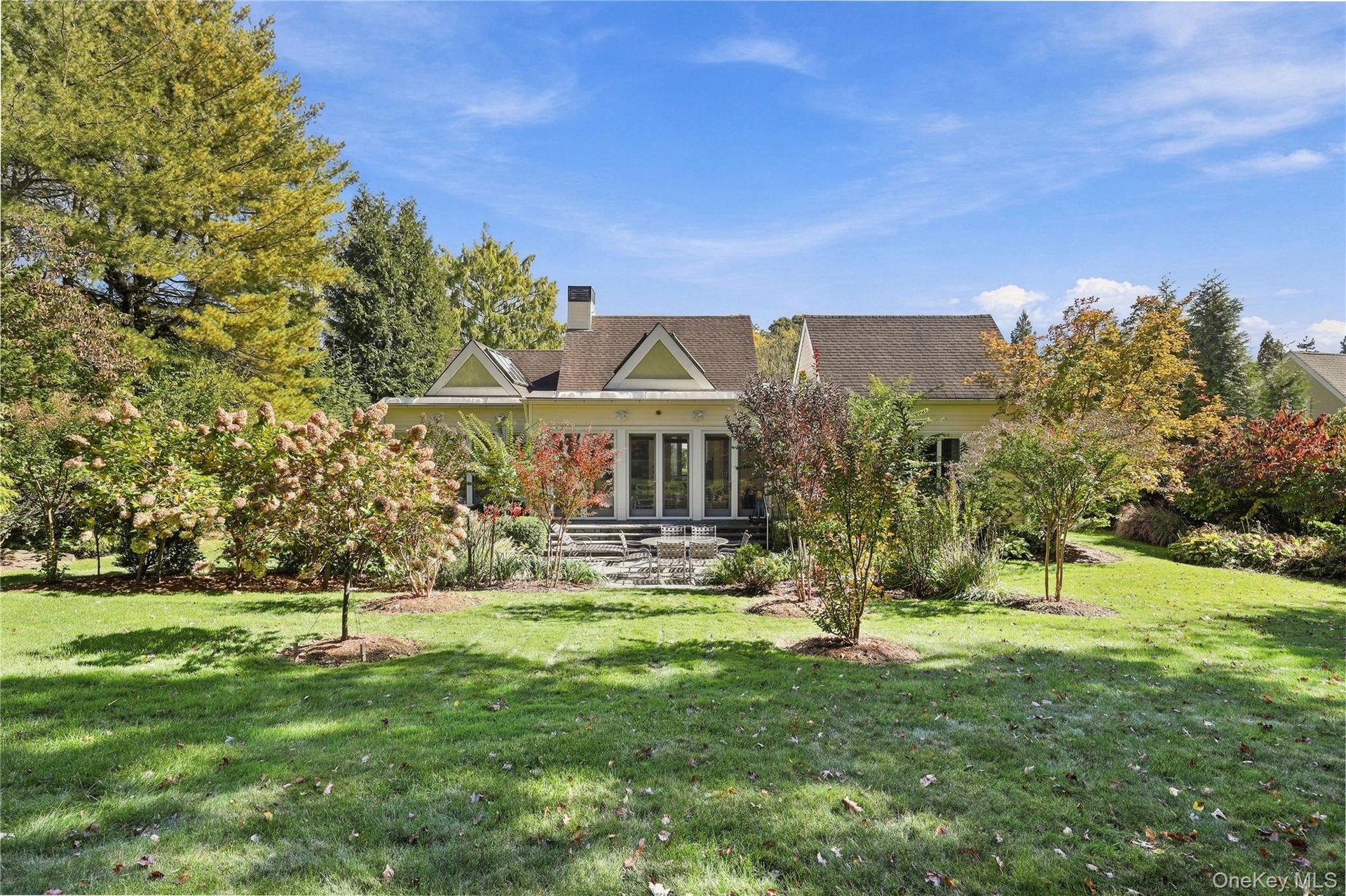 Rear exterior featuring a multi-gabled roofline, ground-level stone patio, and glass-paned double doors set amidst expansive landscaped grounds with m