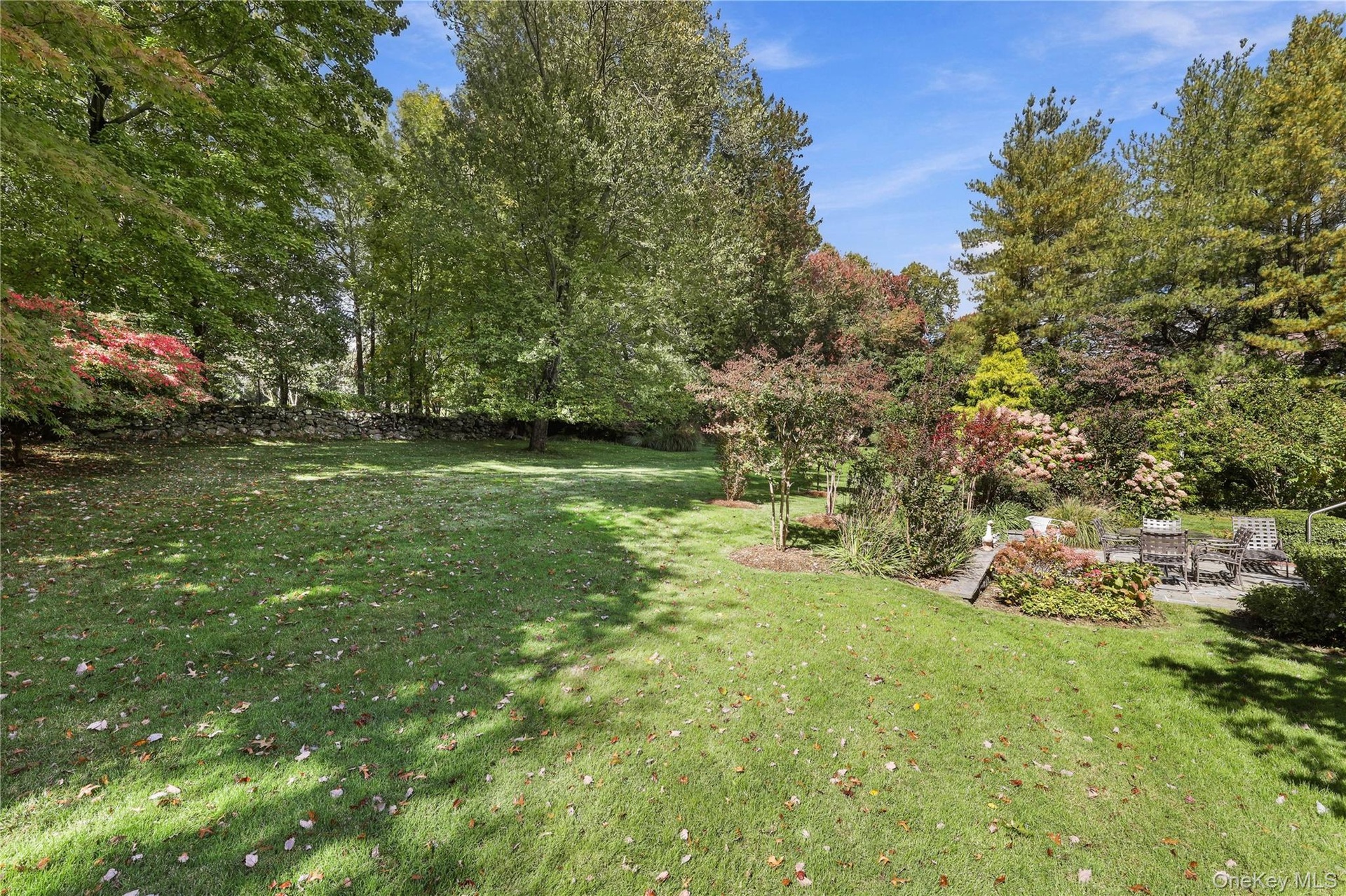 Expansive backyard featuring a manicured lawn, mature trees, a natural stone wall boundary, and varied perennial landscaping
