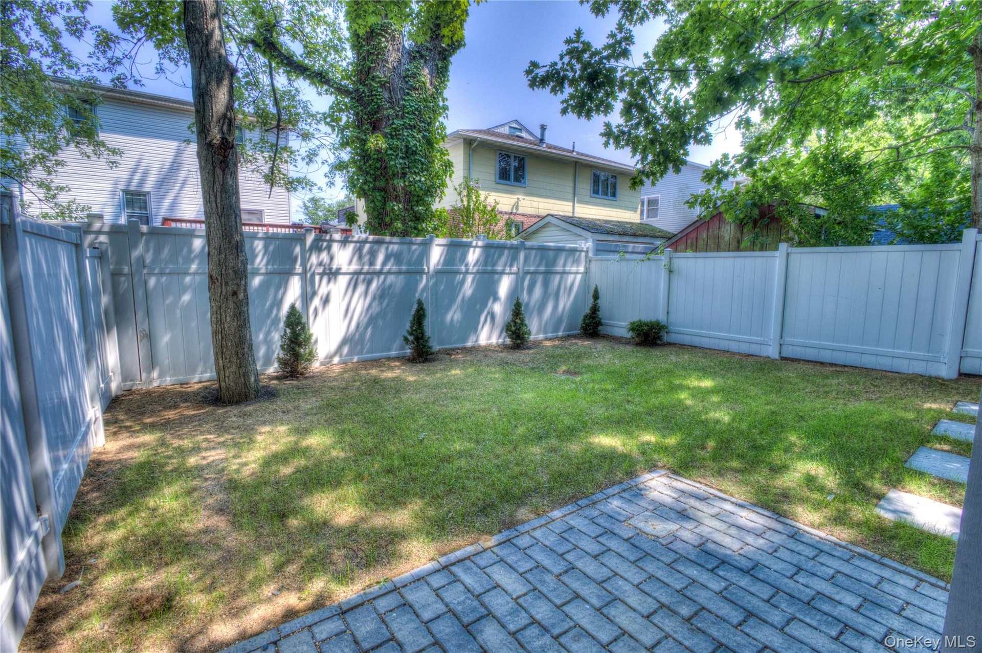 Private backyard featuring a paved stone patio, mature trees, manicured lawn, and full-perimeter vinyl fencing