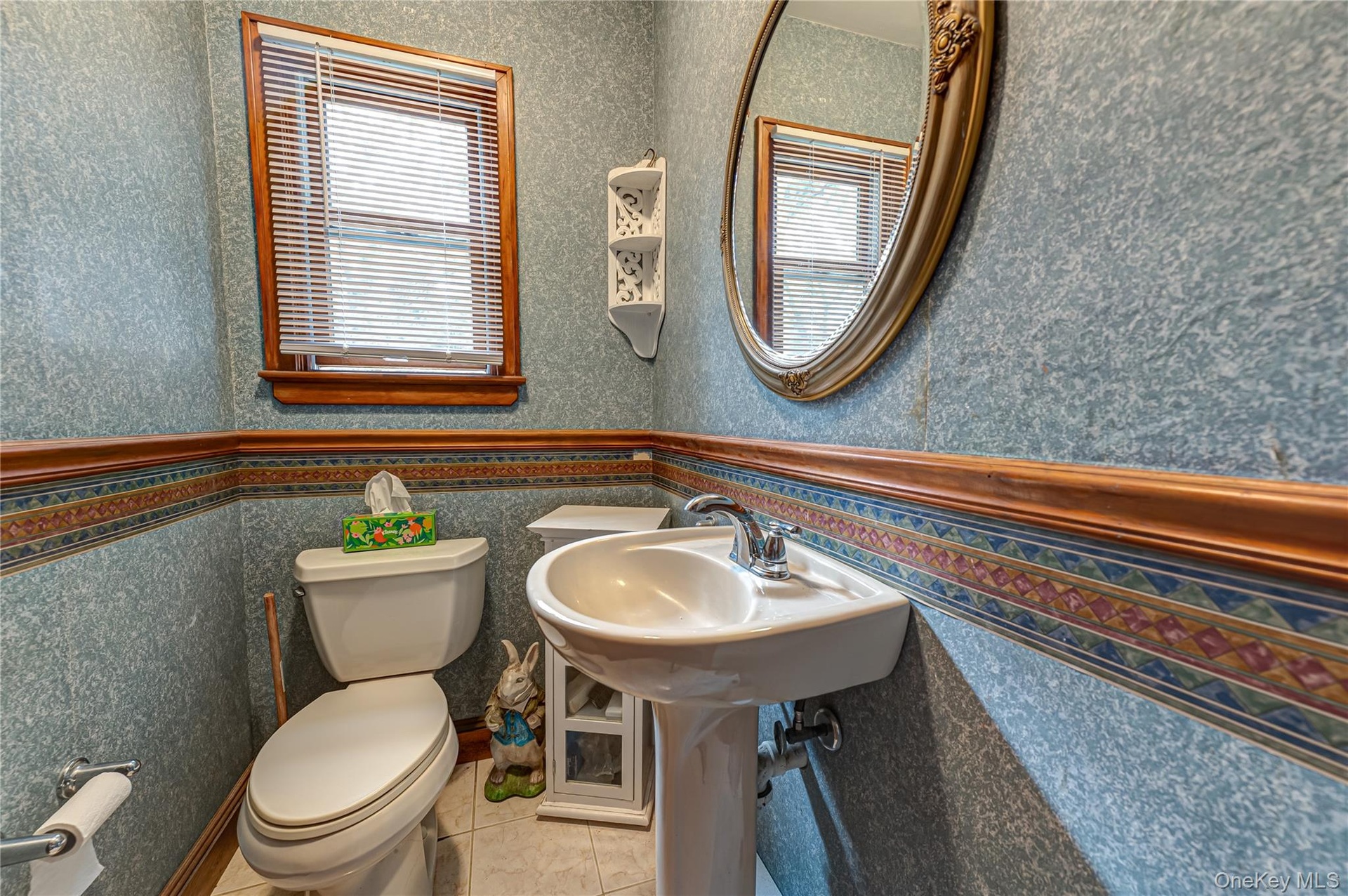 Powder room/half bath with Pedestal sink with chrome faucet, oval wall-mounted mirror, wood-trimmed window.