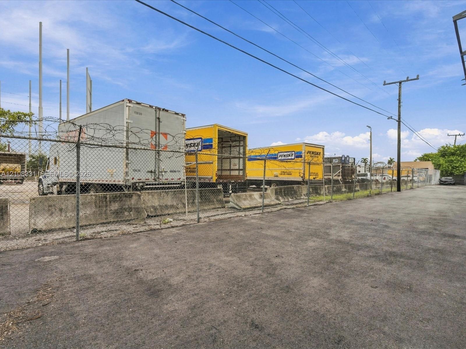 Paved industrial exterior featuring concrete barrier wall, chain-link perimeter fencing, and barbed wire topping