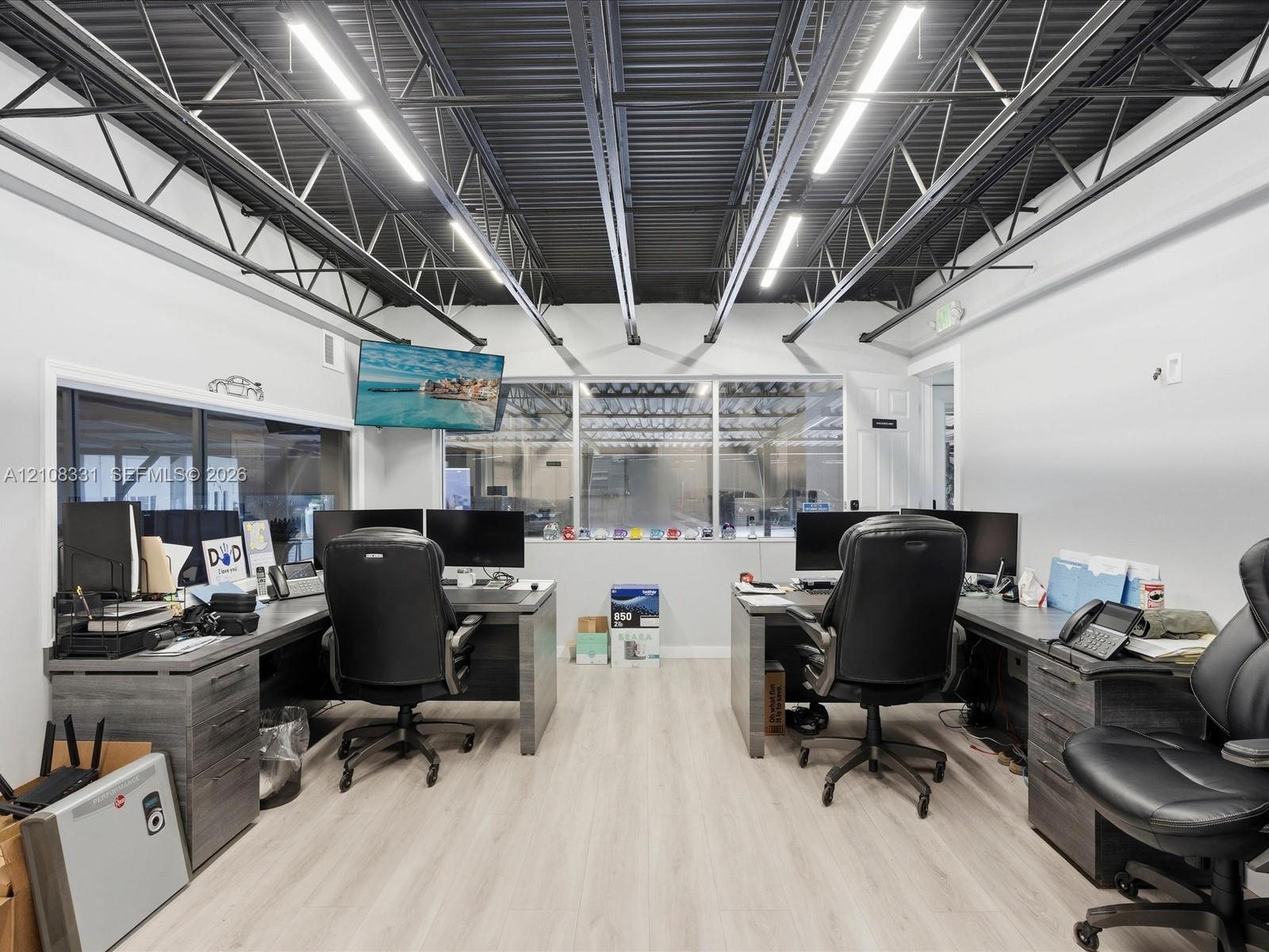 Office space featuring an exposed truss ceiling, industrial-style lighting, neutral wood-finish flooring, and integrated windows providing visibility 