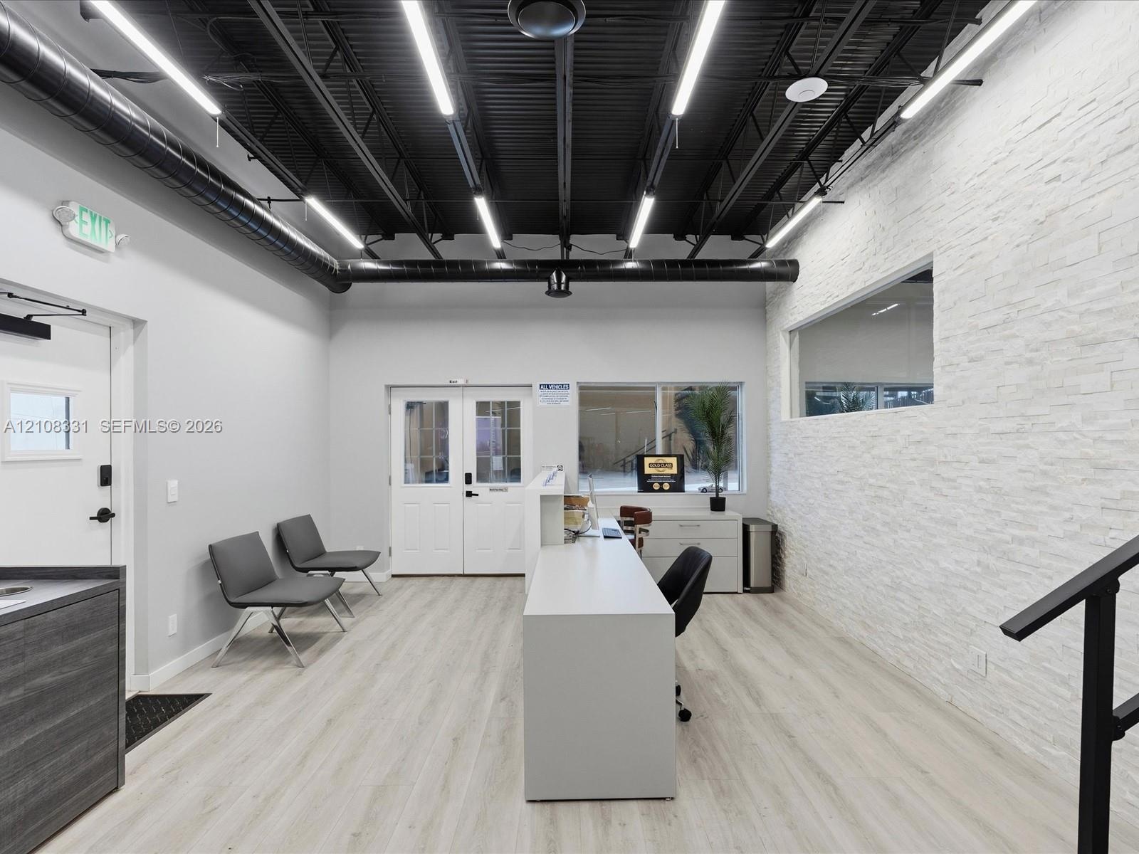 Open-concept interior featuring a high, black exposed-joist ceiling, white stacked-stone accent wall, wood-finish flooring, linear LED lighting, and a