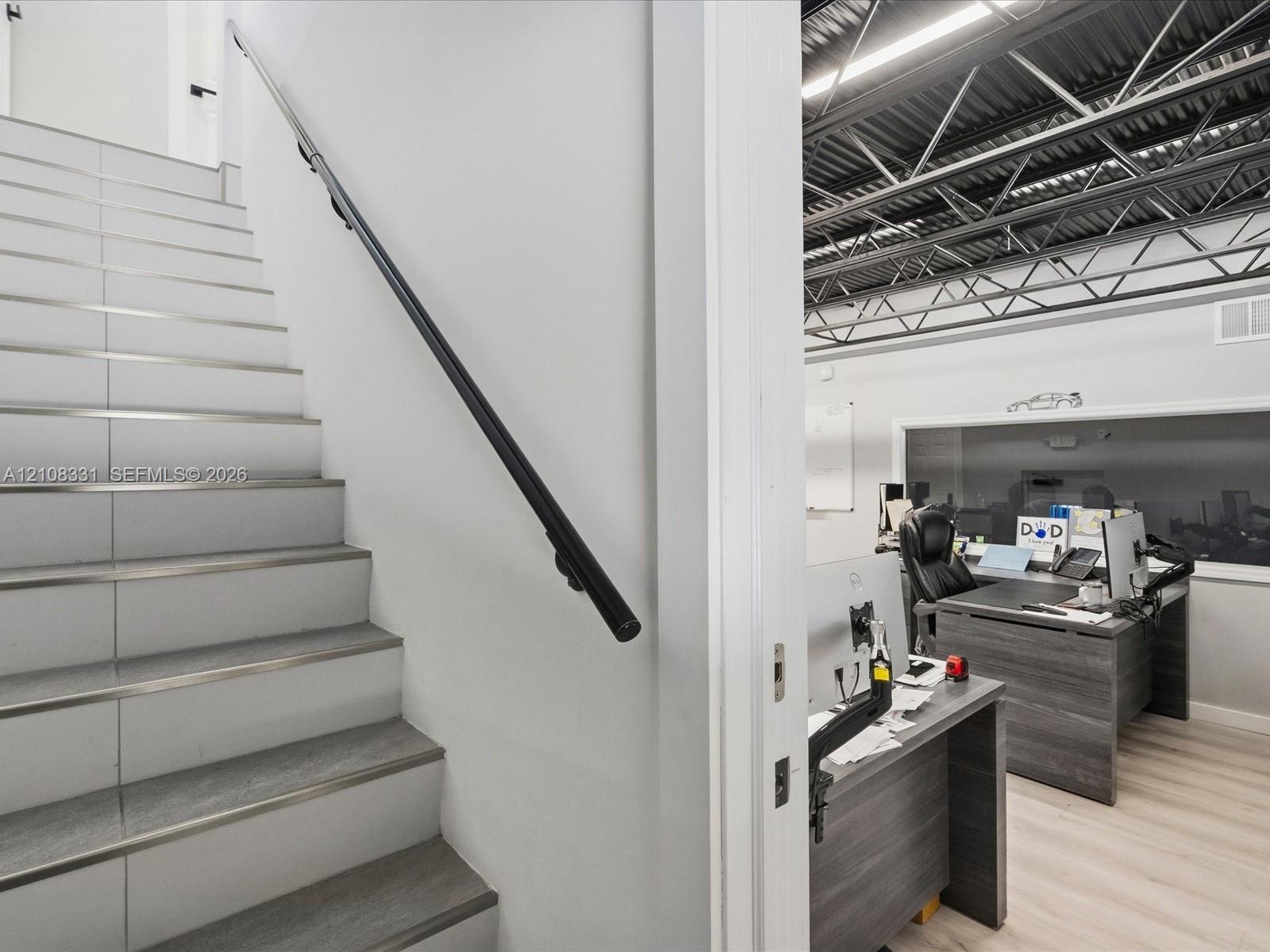 Staircase with wood-finish treads and metal trim, sleek black handrail, and open-plan area featuring exposed industrial ceiling trusses