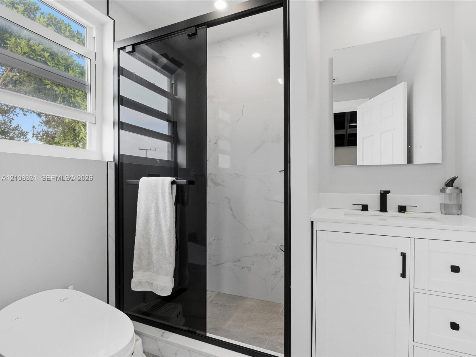 Bathroom featuring a shower with a black-framed glass enclosure, light-toned stone-finish tile surround, white vanity with matte black hardware, and a