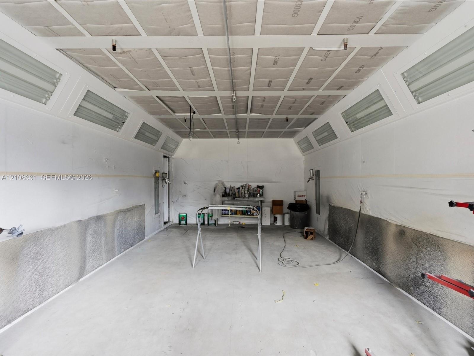 Enclosed professional paint booth featuring integrated overhead fluorescent lighting, paneled ceiling, and concrete flooring
