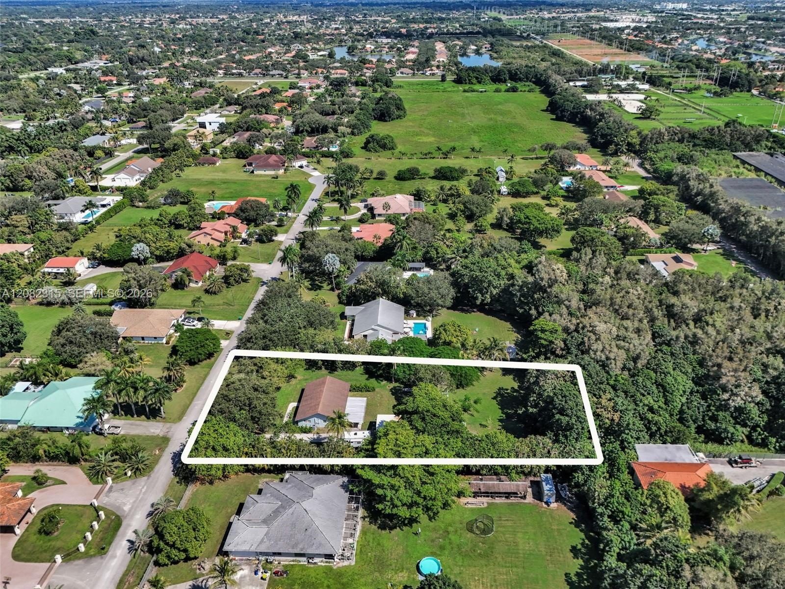 Aerial view of a sprawling rectangular lot featuring extensive open green space, dense perimeter foliage, mature trees, and a single-story structure