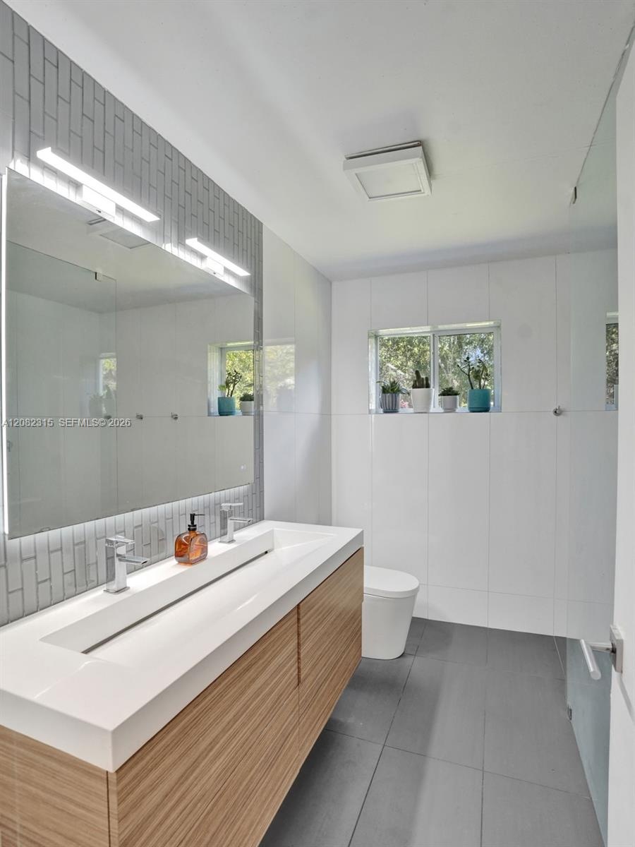 Floating vanity with a double-basin integrated countertop, vertical tile backsplash, full-width vanity mirror, and large-format floor tiling