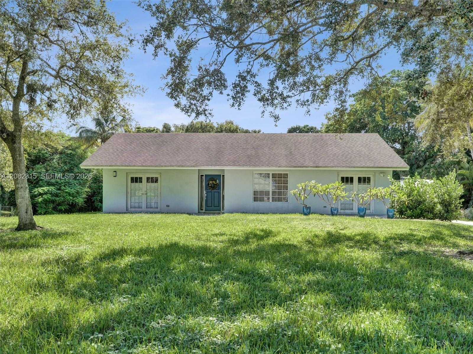 Single-story exterior featuring a light-colored stucco facade, dual glass-paned entry doors, a pitched shingle roof, and an expansive lawn shaded by m
