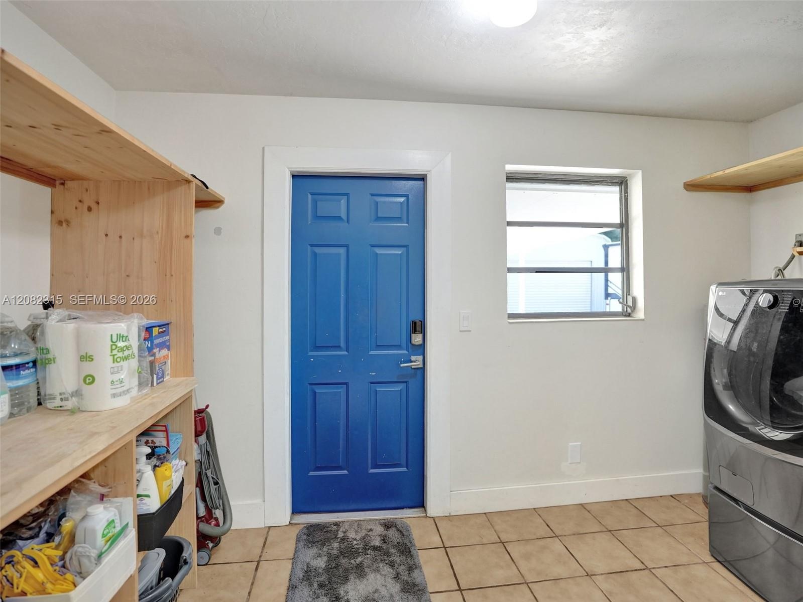 Utility area featuring natural wood-finish open shelving, paneled door with hardware, square ceramic floor tile, and a multi-pane window