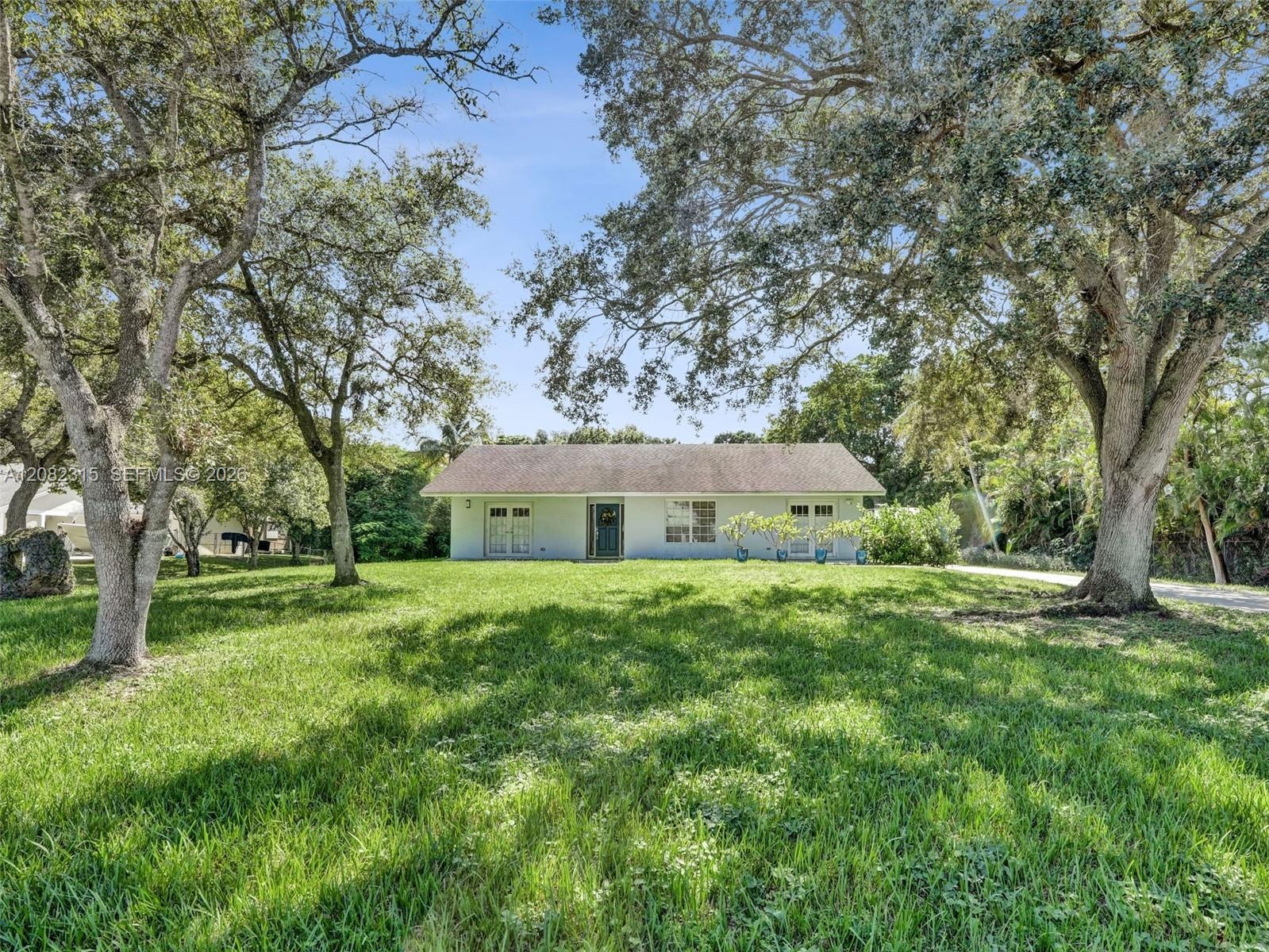 Single-story residence featuring a light-colored stucco exterior, a gable roof, and large mature shade trees situated on a spacious grass lawn