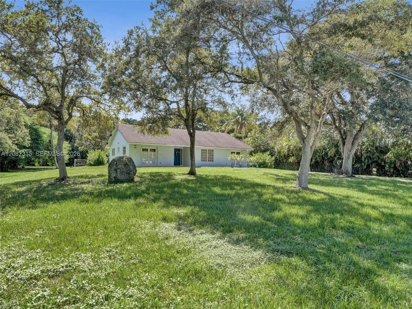 Single-story exterior featuring light-colored siding, a gabled roof, mature oak trees, and expansive lawn space