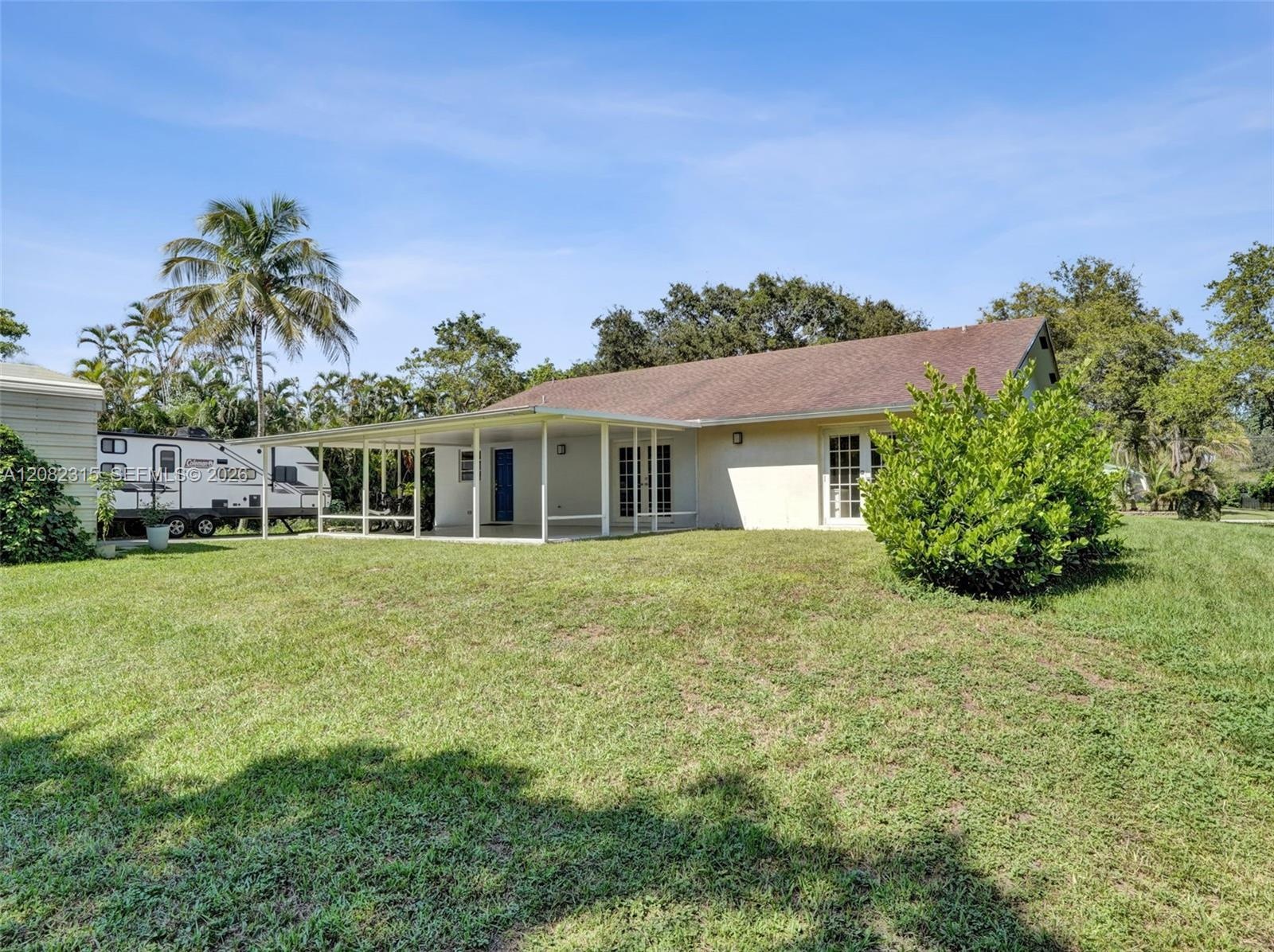 Single-story residence featuring a covered patio, shingle roof, exterior lighting fixtures, and a spacious grass yard with mature vegetation