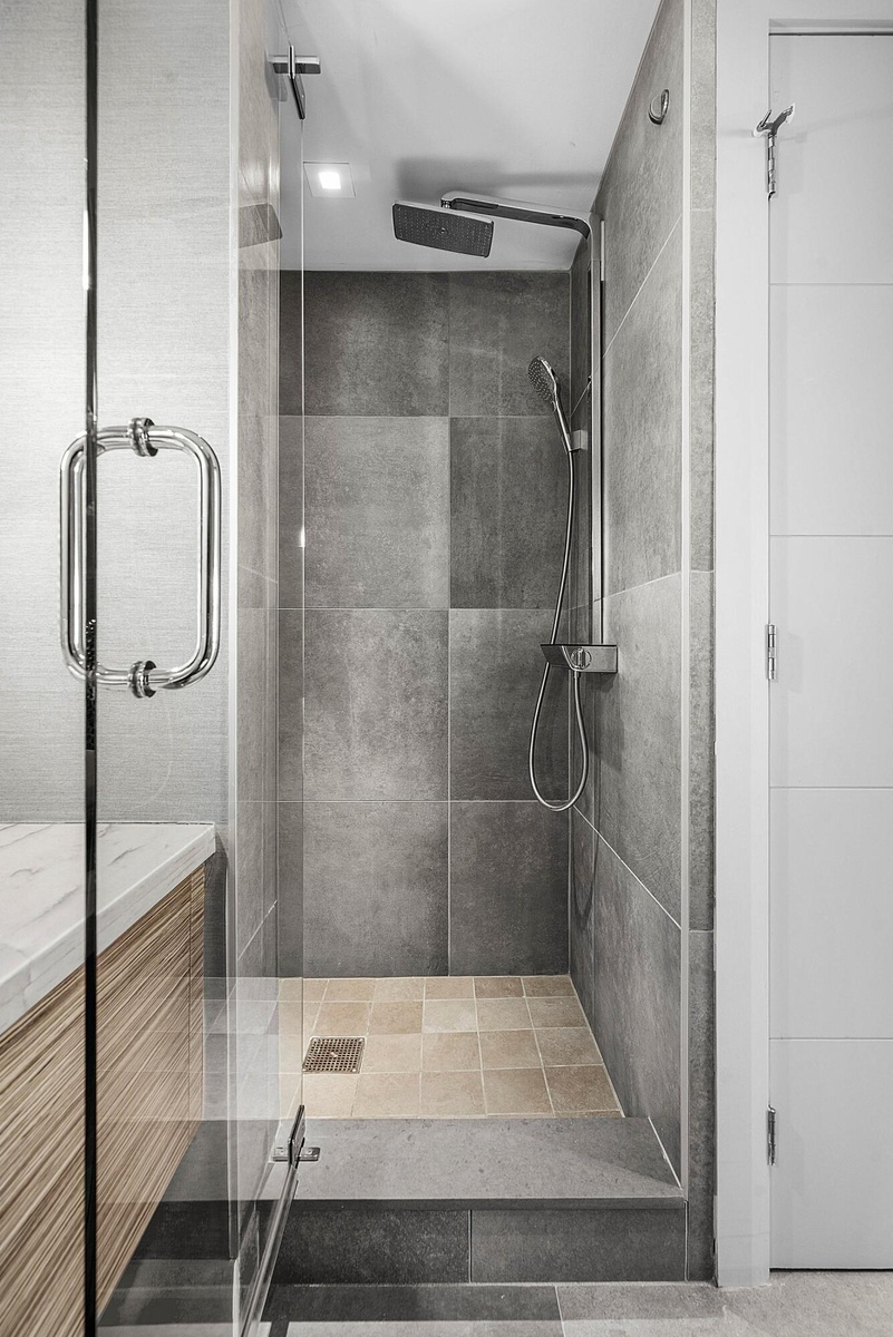 Walk-in shower featuring large-format gray tile walls, a dedicated rainfall showerhead, handheld sprayer, glass enclosure, and a stone-tiled floor