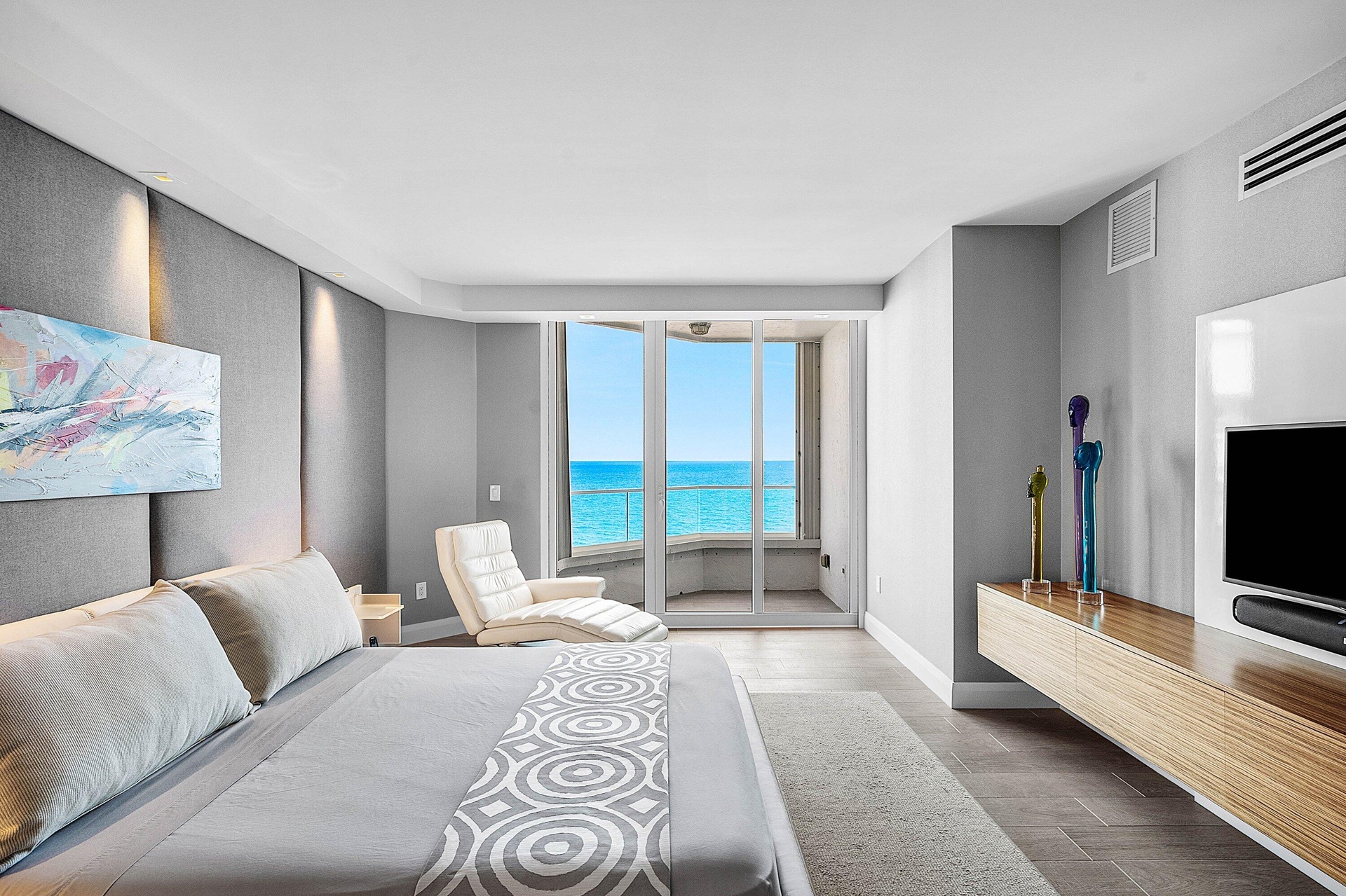 Floor-to-ceiling glass sliding doors opening to a balcony, wall-to-wall upholstered headboard paneling, wall-mounted wood-finish media console, and la