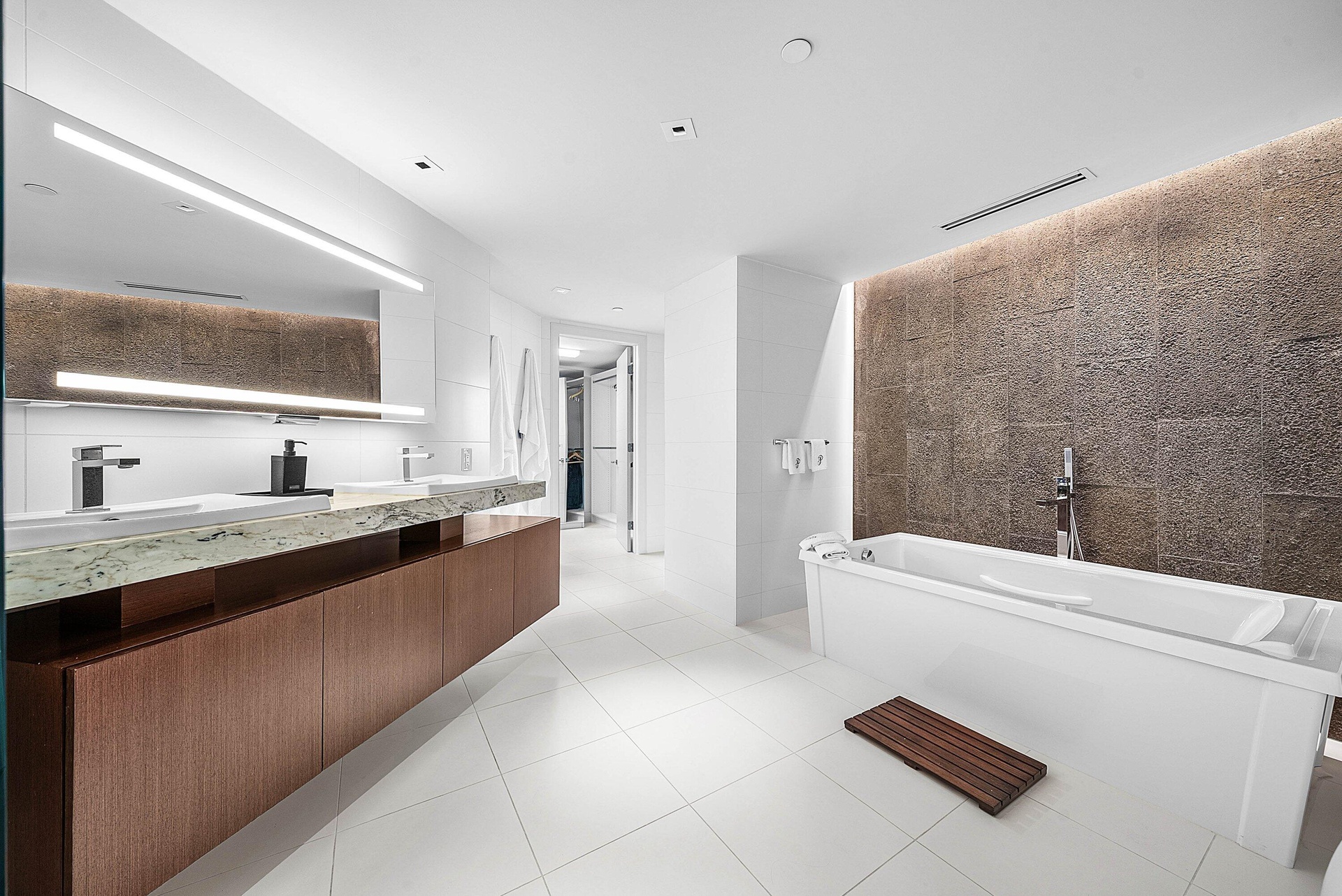Floating wood-finish double vanity with stone countertop, large LED-backlit mirror, soaking tub, textured stone feature wall, and recessed ceiling lig
