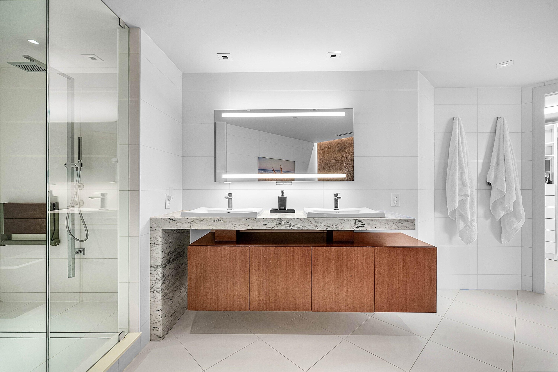 Floating wood-finish vanity with dual integrated sinks and stone-surfaced waterfall countertop, complemented by an illuminated vanity mirror and glass