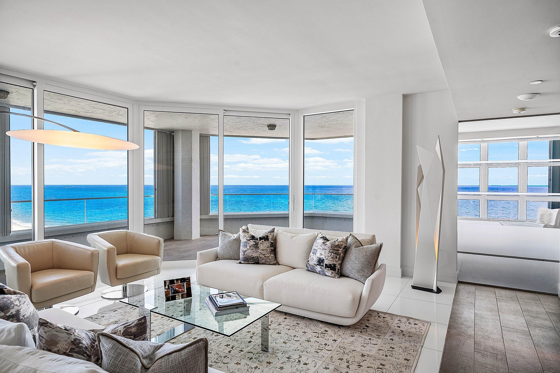 Floor-to-ceiling panoramic windows, expansive balcony with glass railings, polished tile flooring, recessed lighting, and coastal water backdrop