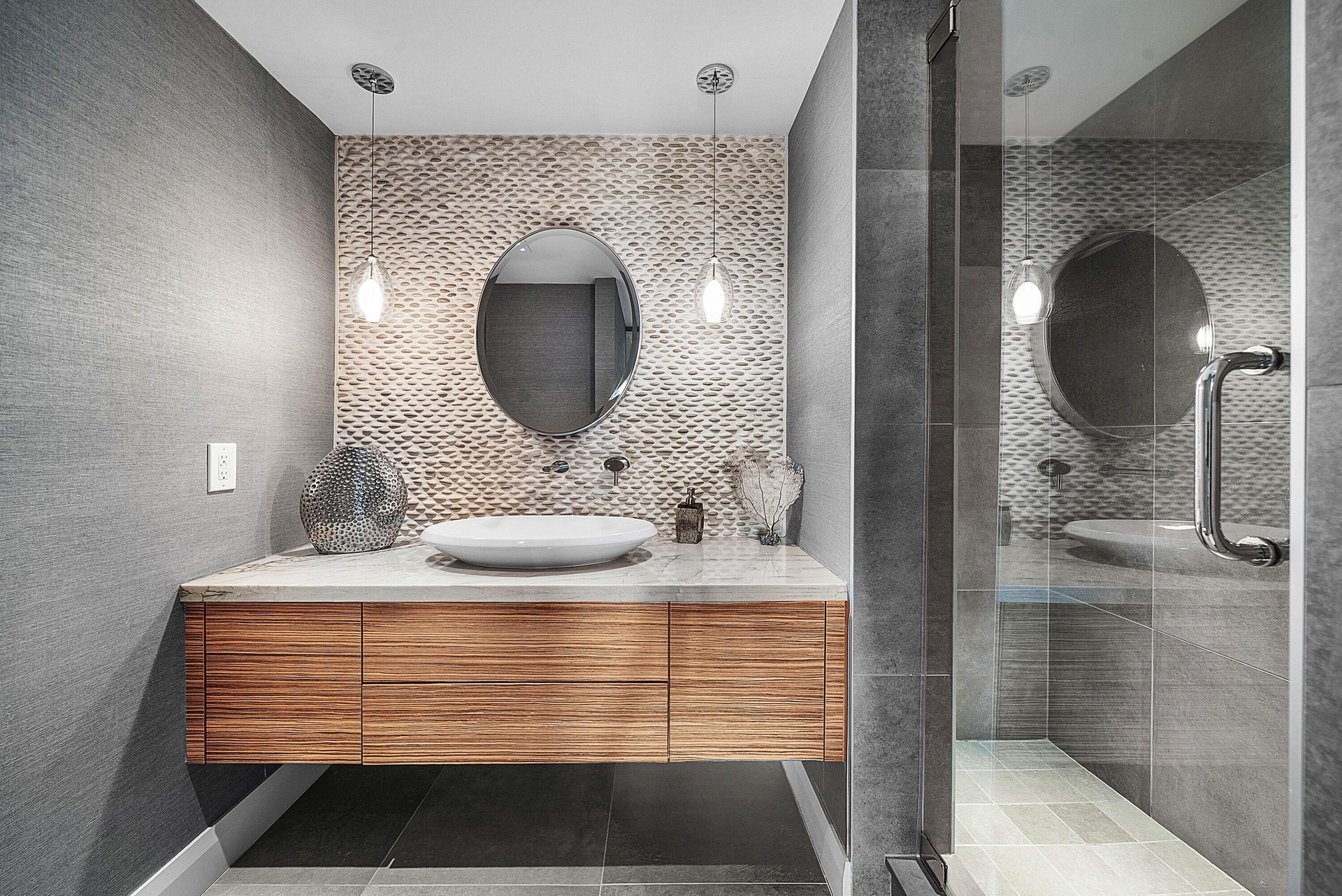 Floating wood-finish vanity with a vessel sink and stone countertop, pebble-texture accent wall, dual pendant lighting, and a glass-enclosed shower wi
