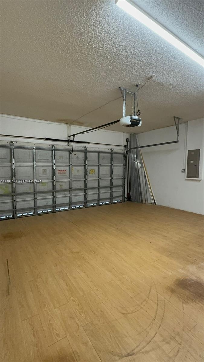 Enclosed garage featuring wood-finish flooring, metal sectional overhead door, ceiling-mounted automatic door opener, and integrated electrical servic