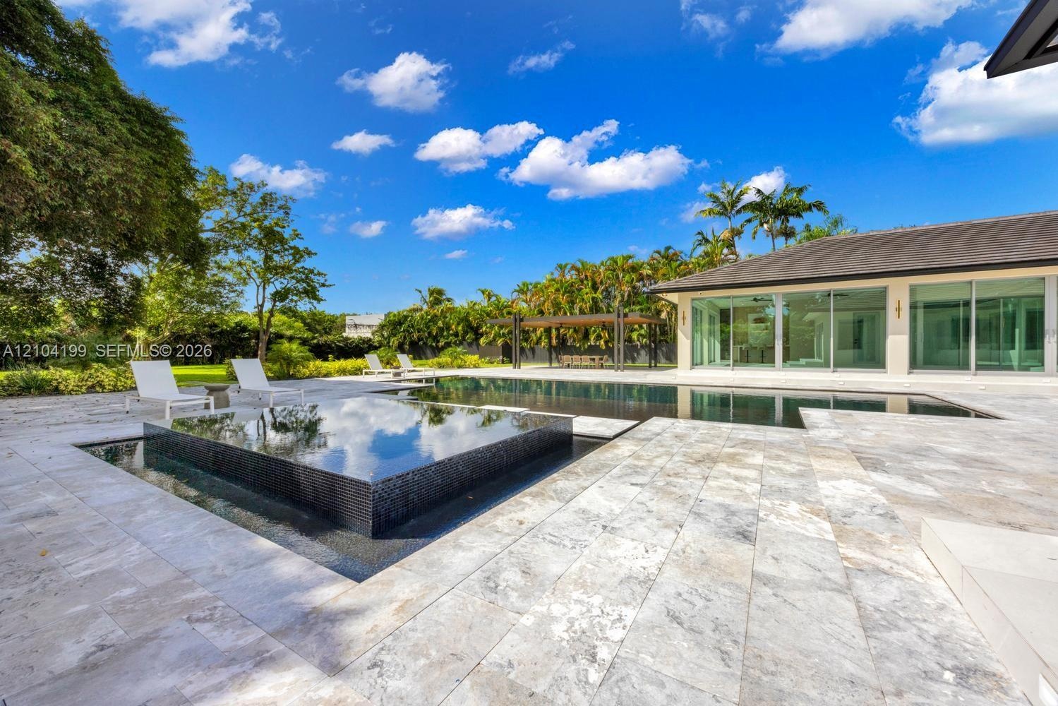 Backyard featuring a custom infinity-edge pool, a raised square spa with tile surfacing, extensive stone paver decking, and a covered pavilion structu
