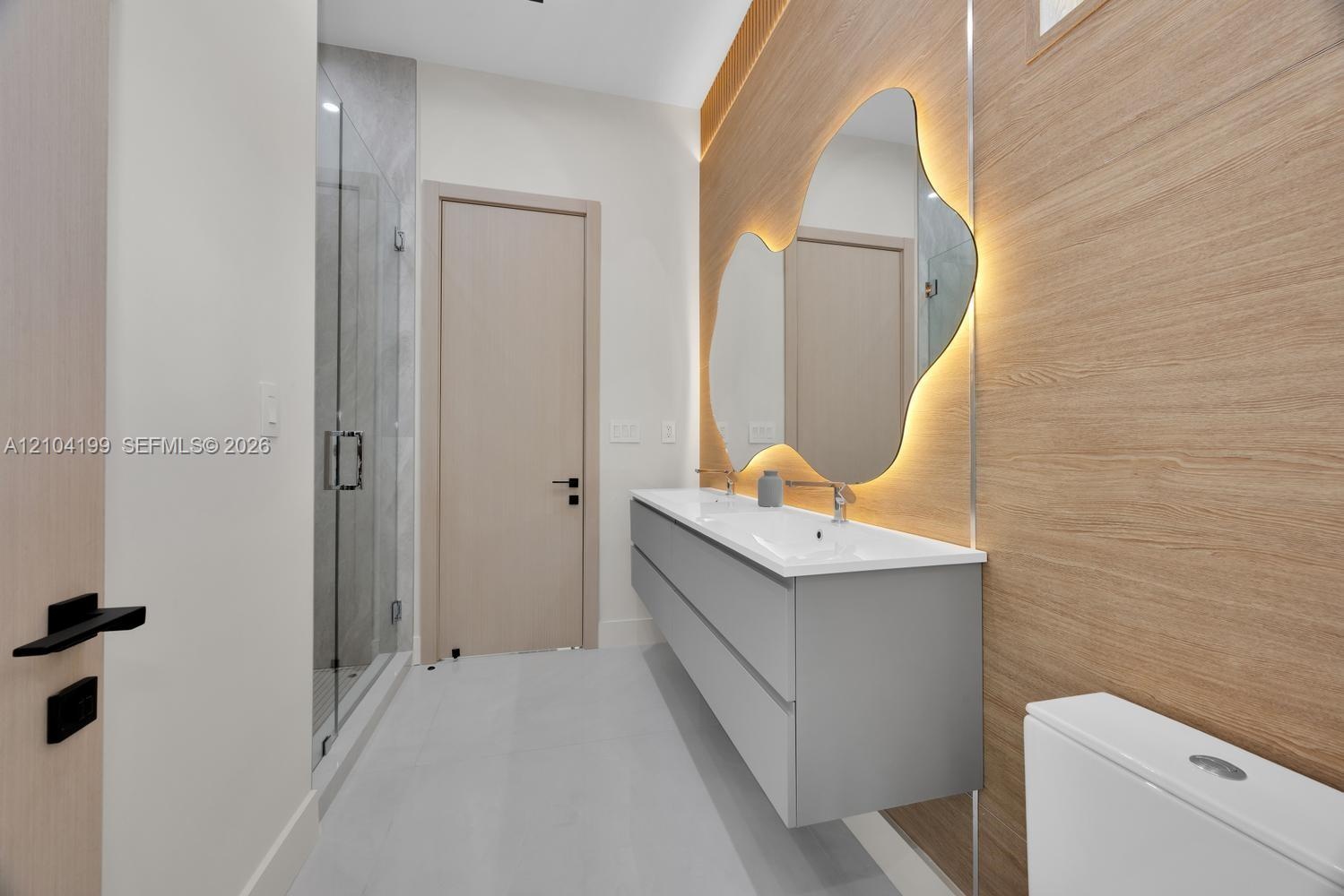 Floating grey vanity with integrated double sinks, oversized organic-shaped wall mirror with backlighting, wood-finish wall paneling, and glass-enclos