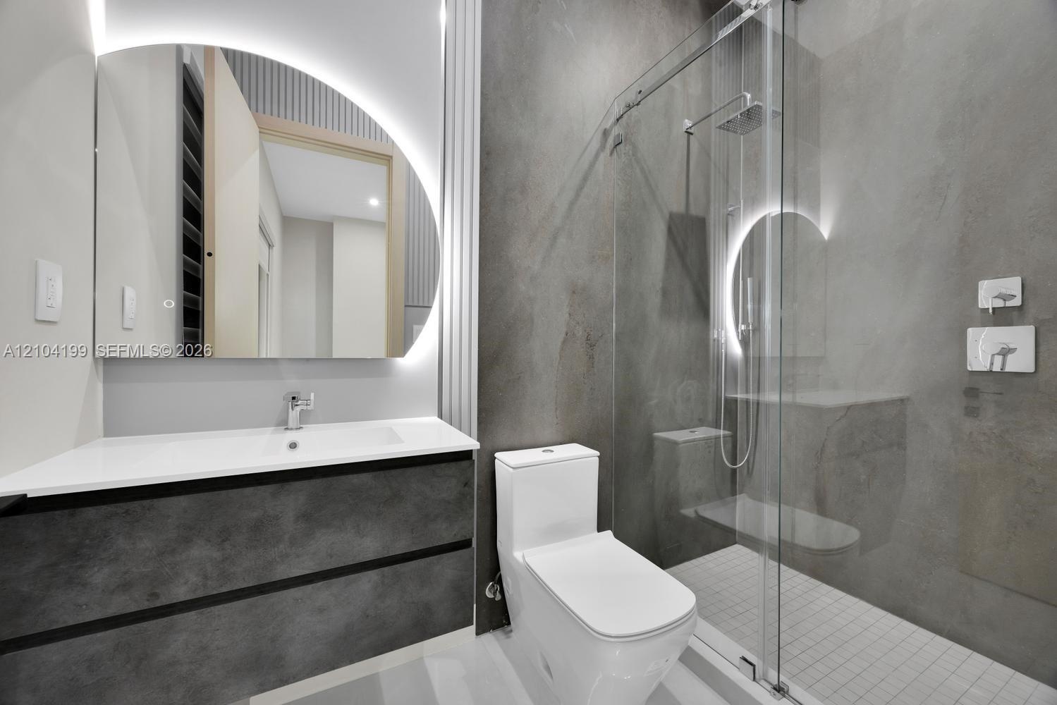 Bathroom featuring a floating vanity with a white countertop, an arched LED-backlit mirror, large-format grey tile walling, and a walk-in shower with 