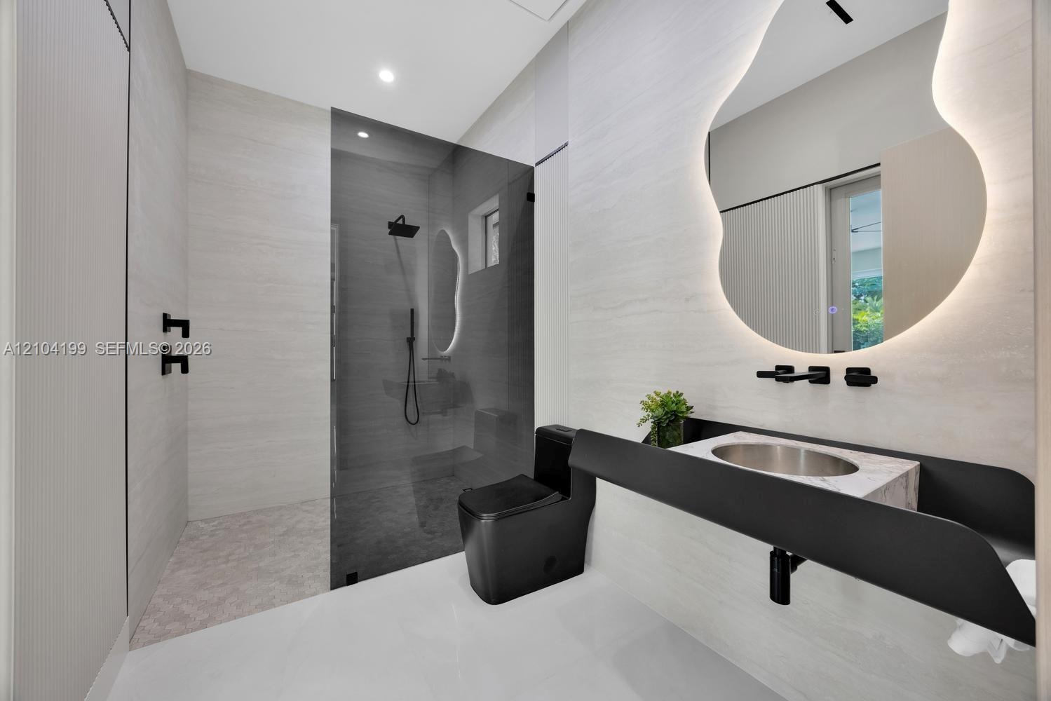 Bathroom featuring a wall-mounted floating vanity with a stone countertop, backlit organic-shaped mirror, matte black fixtures, and a walk-in shower w