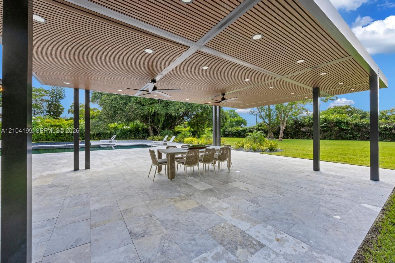 Covered outdoor patio featuring a slatted wood-finish ceiling, recessed lighting, ceiling fans, stone paver flooring, and a swimming pool