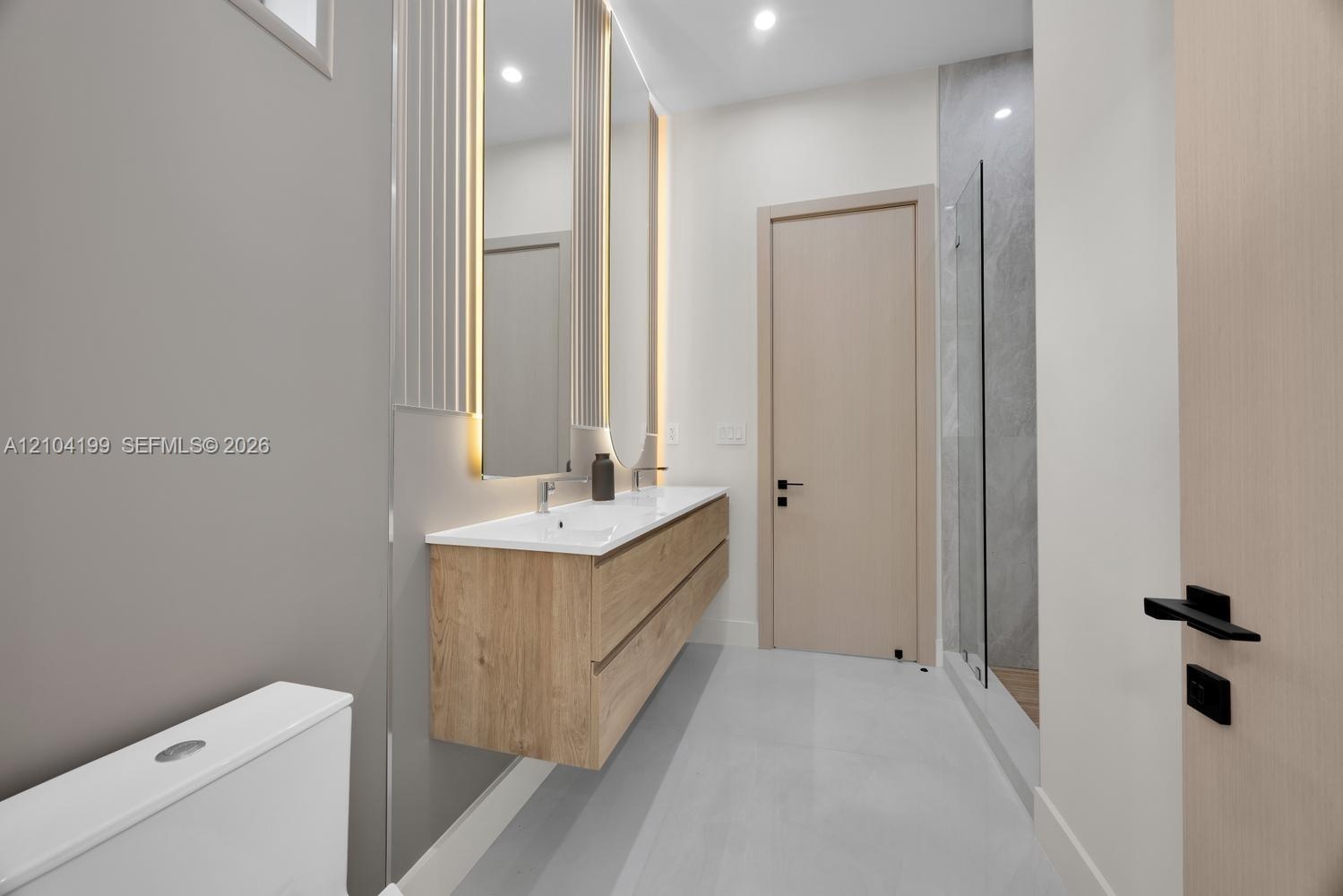 Floating wood-finish double vanity with integrated sinks, dual backlit mirrors, matte black hardware, and a walk-in shower with large-format tile surr