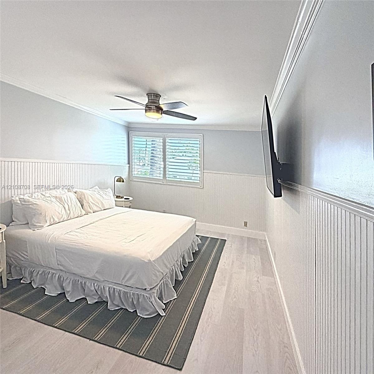 Room featuring wood-finish flooring, white beadboard wainscoting, crown molding, plantation shutters, and a central ceiling fan with an integrated lig