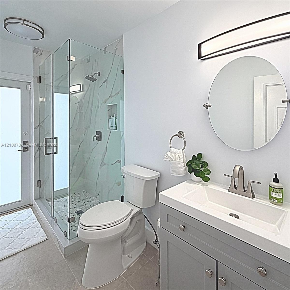 Glass-enclosed shower with stone-finish wall tile and pebble-texture flooring, grey shaker-style vanity with integrated sink, and circular wall-mounte