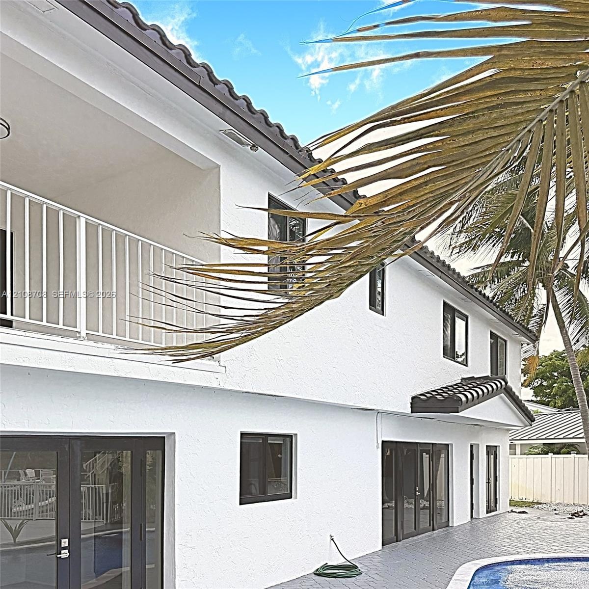 Exterior facade featuring white stucco finish, dual-level covered balcony with metal railing, tile roof, dark-frame glass doors, and a paved pool surr