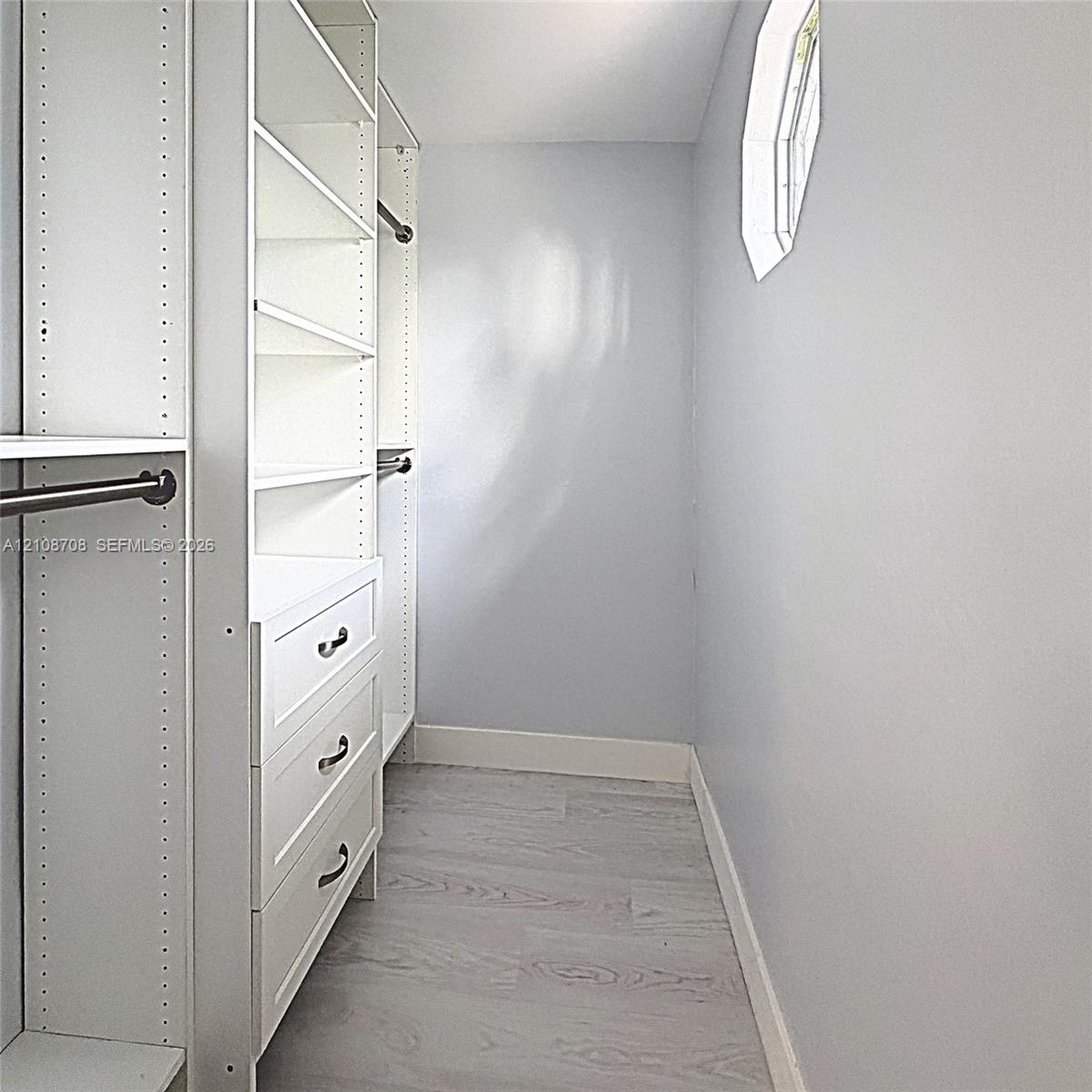 Walk-in closet featuring custom white built-in shelving, integrated three-drawer storage, adjustable garment rods, wood-finish flooring, and a small h