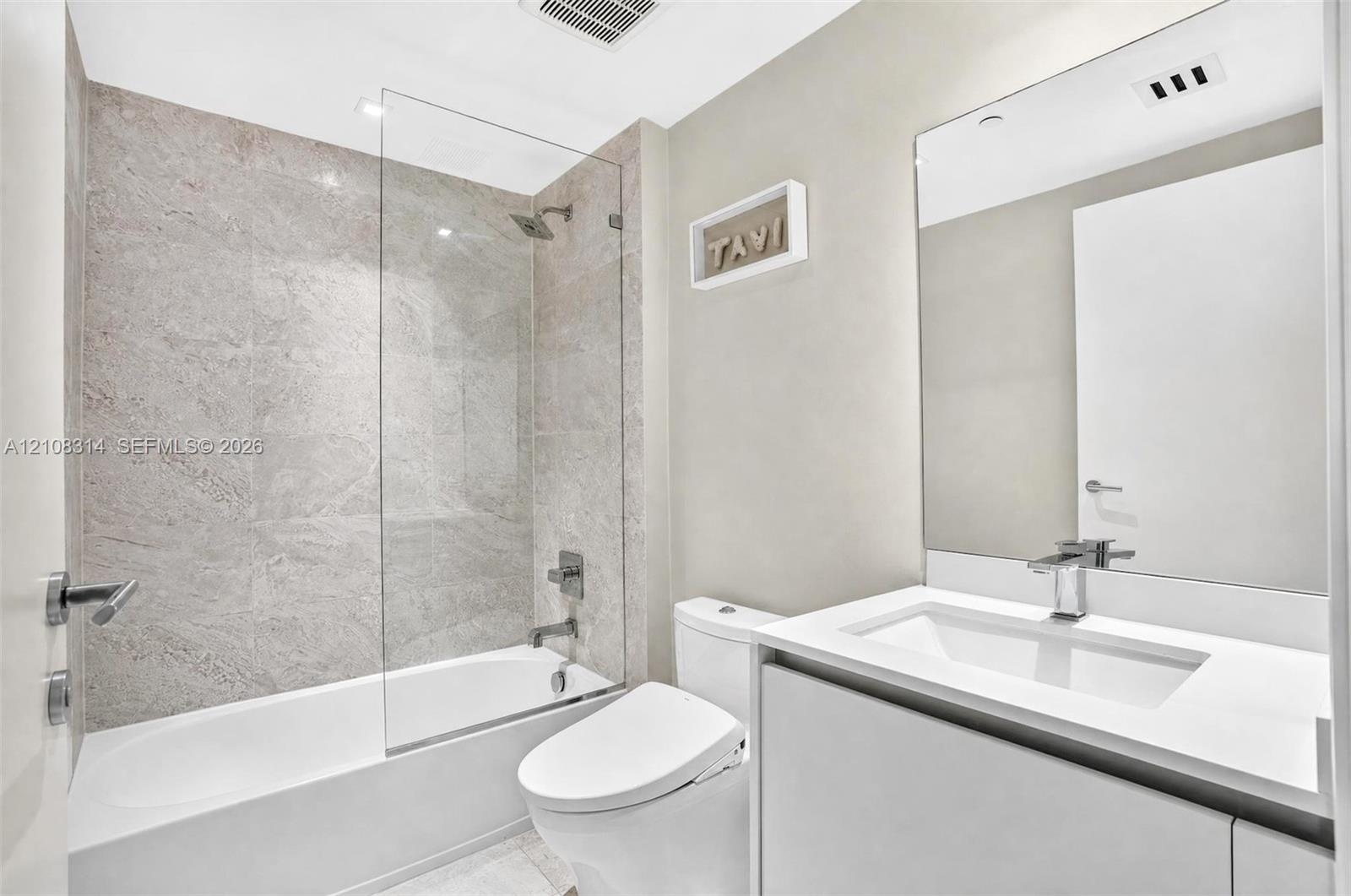 Bathroom featuring a vanity with a white countertop and integrated sink, frameless glass shower enclosure, stone-finish tile surround, and large wall-