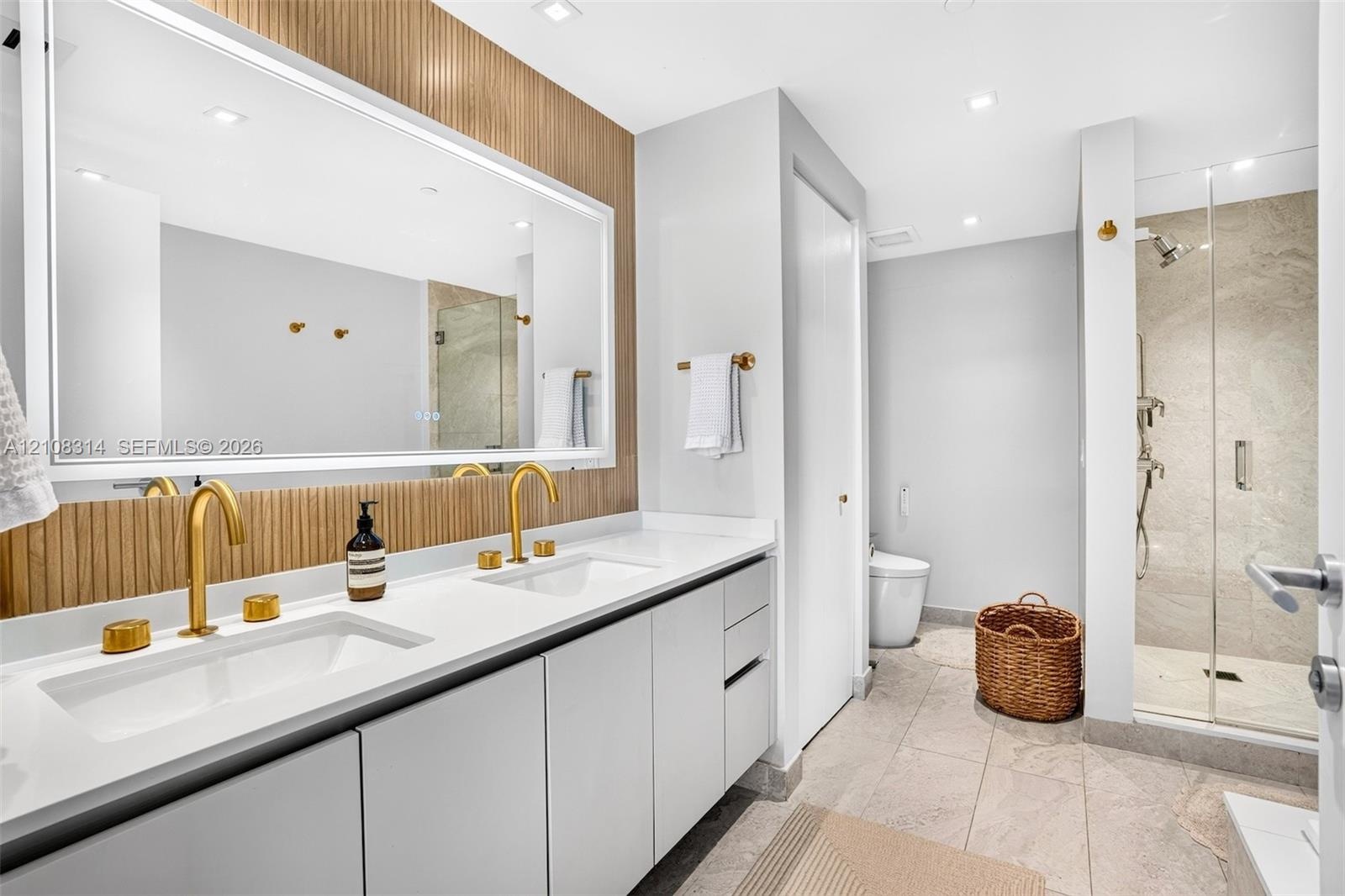 Dual-sink vanity with white surface, metallic gold-finish faucets, large-scale vanity mirror with integrated lighting, wood-slat wall paneling, and wa