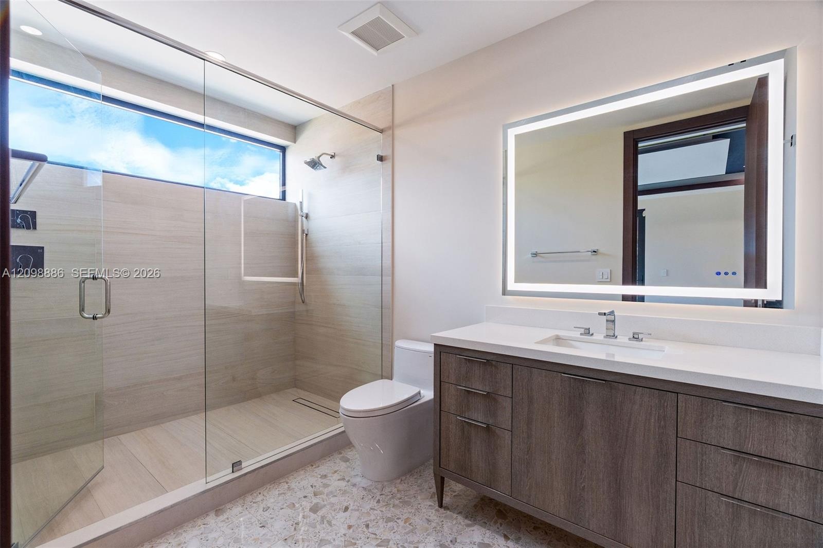Bathroom featuring a walk-in shower with large-format tile surround, frameless glass enclosure, a wood-finish vanity with a stone countertop, and an i