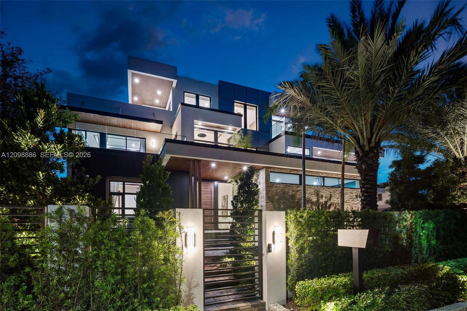 Modern multi-level exterior featuring clean geometric lines, stacked stone accents, recessed ceiling lighting, glass balcony railings, and a gated ent