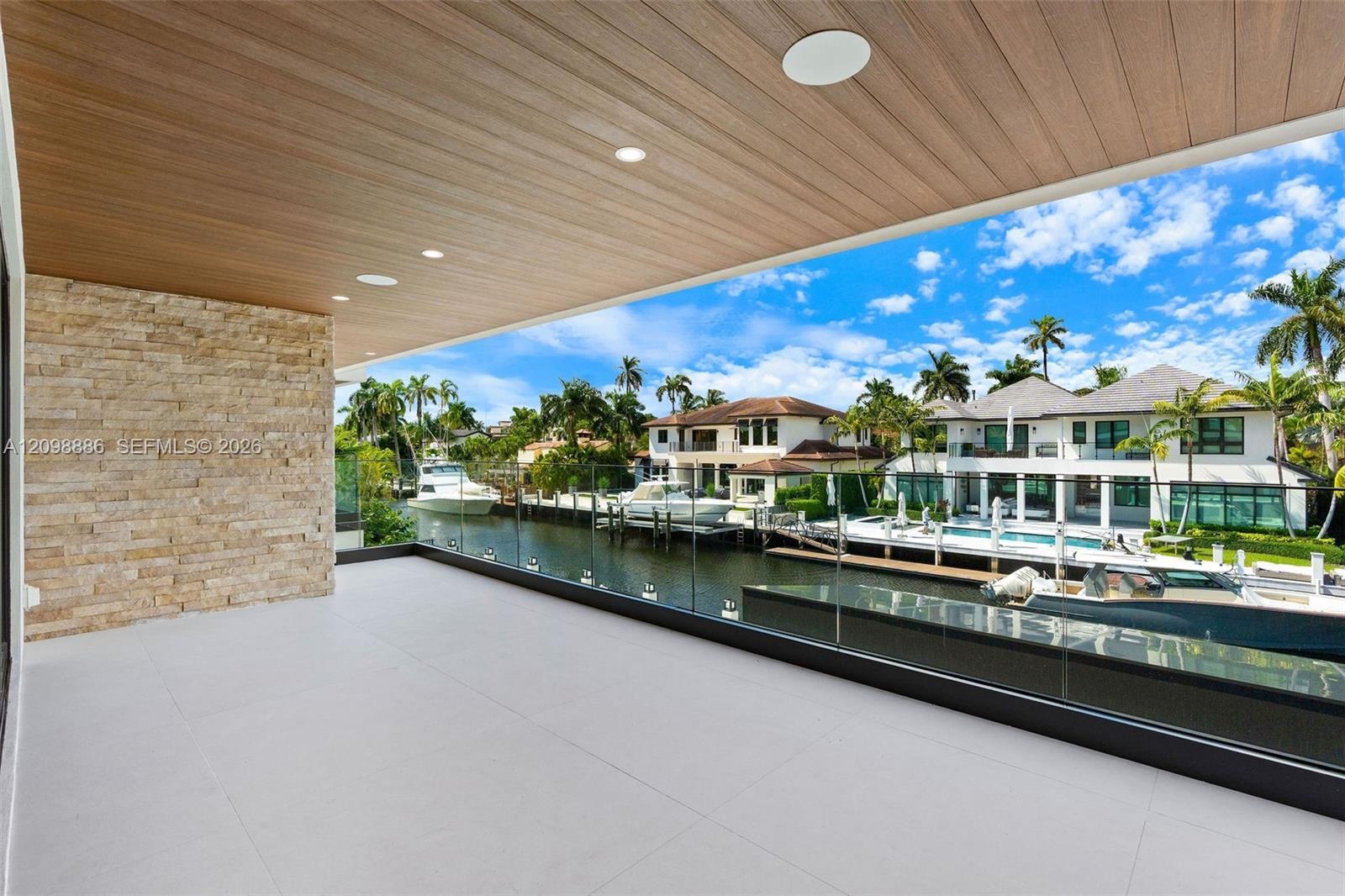 Covered balcony featuring wood-finish ceiling paneling, stacked stone accent wall, large-format tile flooring, and a frameless glass railing overlooki