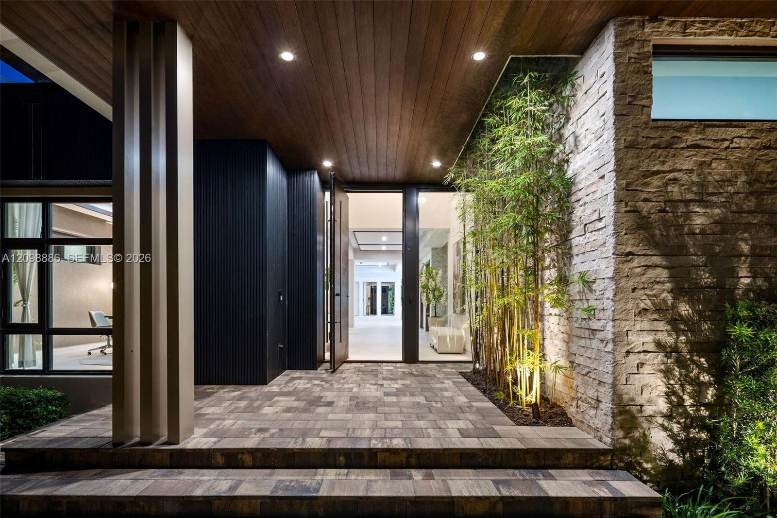 Covered entryway featuring a wood-panelled ceiling, stacked stone accent wall, multi-tiered stone paver stairs, vertical column detailing, and recesse