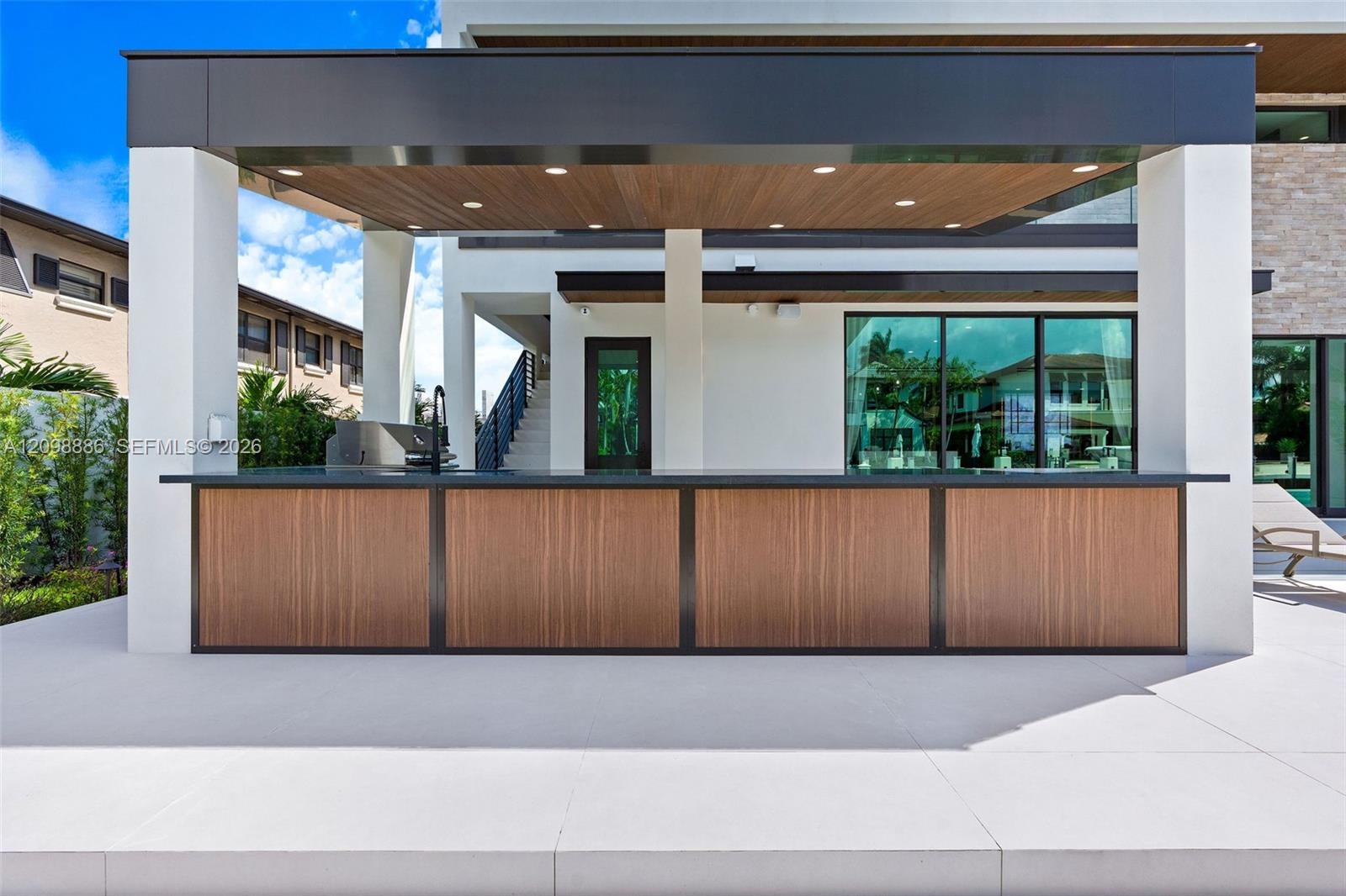 Outdoor kitchen featuring a wood-panel base, dark stone countertop, built-in sink, recessed ceiling lighting, and a metal-framed roof structure