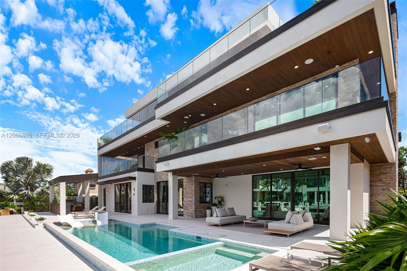 Contemporary multi-level exterior featuring stacked stone masonry, a glass-railed balcony, deep recessed wood-finish soffits, and a rectangular swimmi
