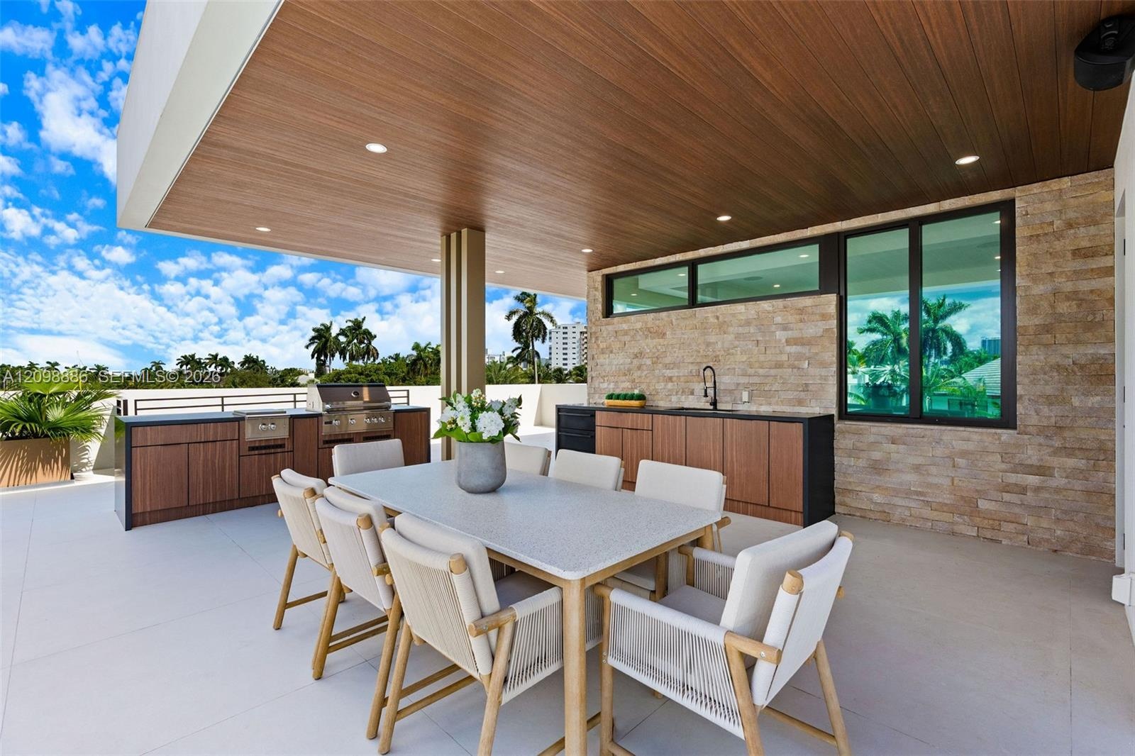 Covered outdoor living space featuring a wood-finish ceiling with recessed lighting, stone veneer wall, integrated kitchenette with stainless steel gr
