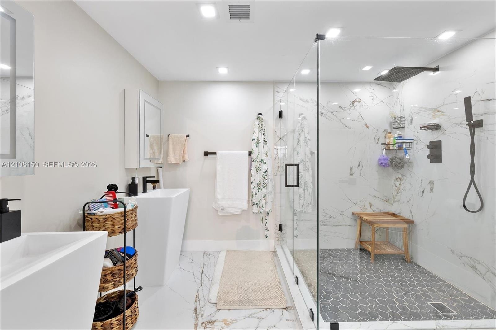 Bathroom featuring large-format stone-finish flooring, pedestal sinks, and a walk-in glass-enclosed shower with contrasting hexagon tile flooring, sto