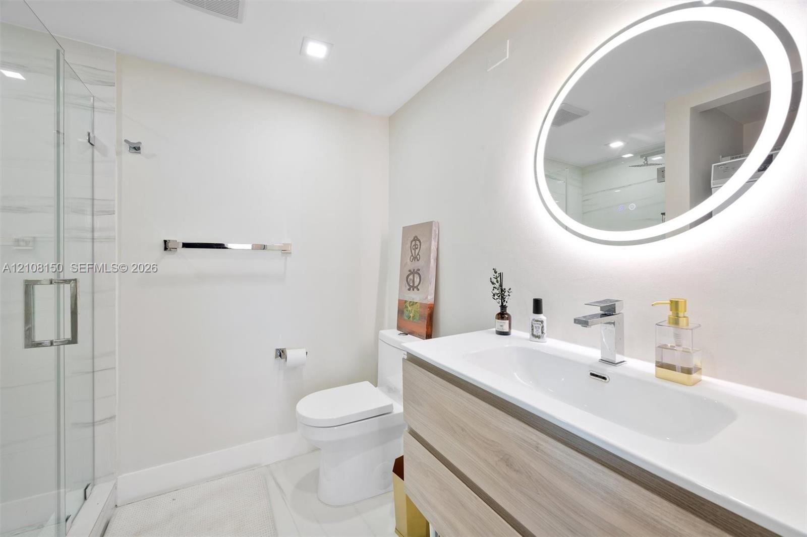 Bathroom featuring a wall-mounted vanity with wood-finish cabinetry, an integrated sink with a chrome faucet, a backlit oval mirror, and a walk-in gla