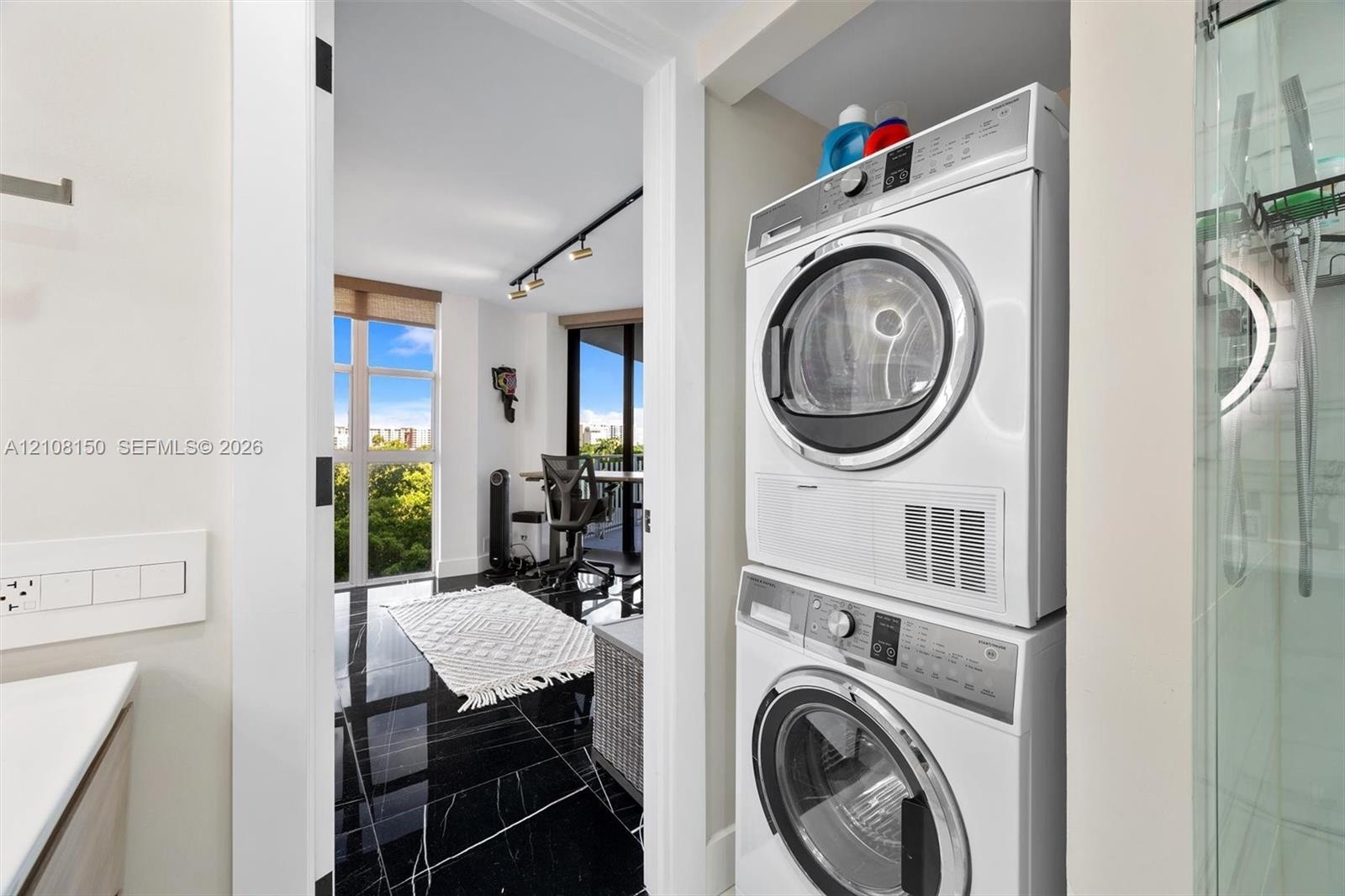 Stacked front-load laundry unit, black polished flooring with white veining, floor-to-ceiling windows, and track lighting