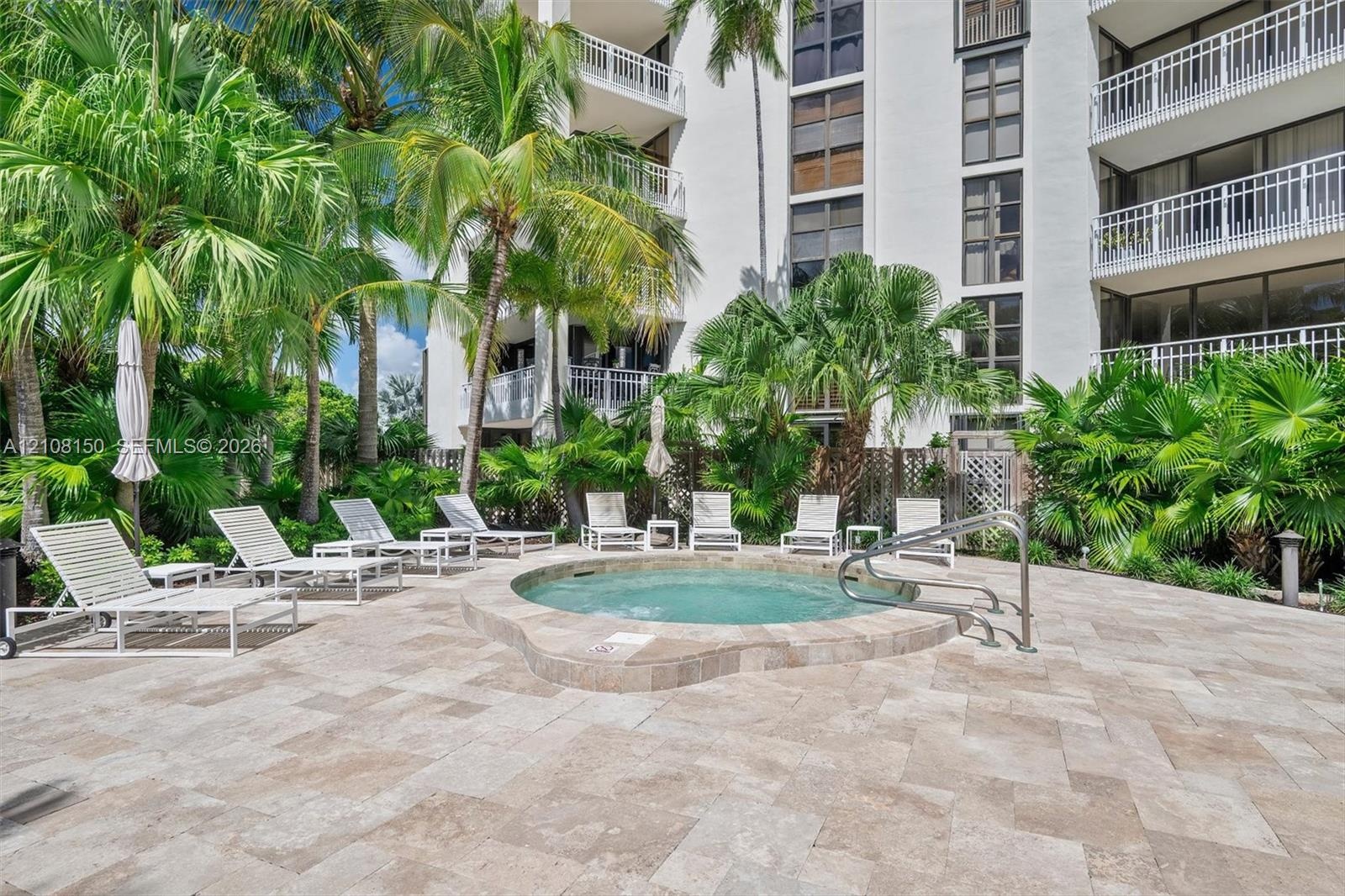 Outdoor spa featuring a stone-tile surround, light-toned paved deck, metal handrail, and tropical landscaping