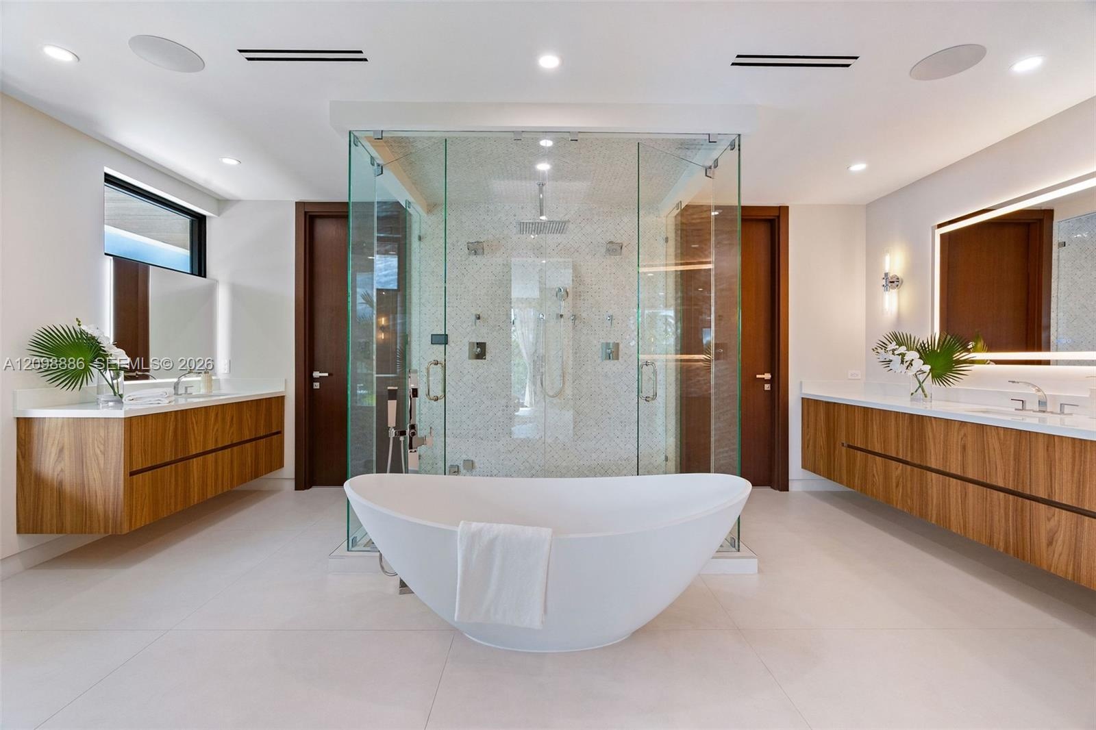 Dual floating wood-finish vanities with rectangular vessel sinks, floor-to-ceiling glass-enclosed shower with tiled interior, and freestanding soaking
